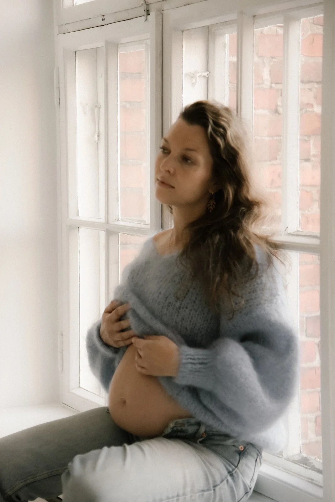 Odotuskuva, jossa raskaana oleva nainen istuu ikkunalaudalla ja pitelee hellästi vatsaansa.