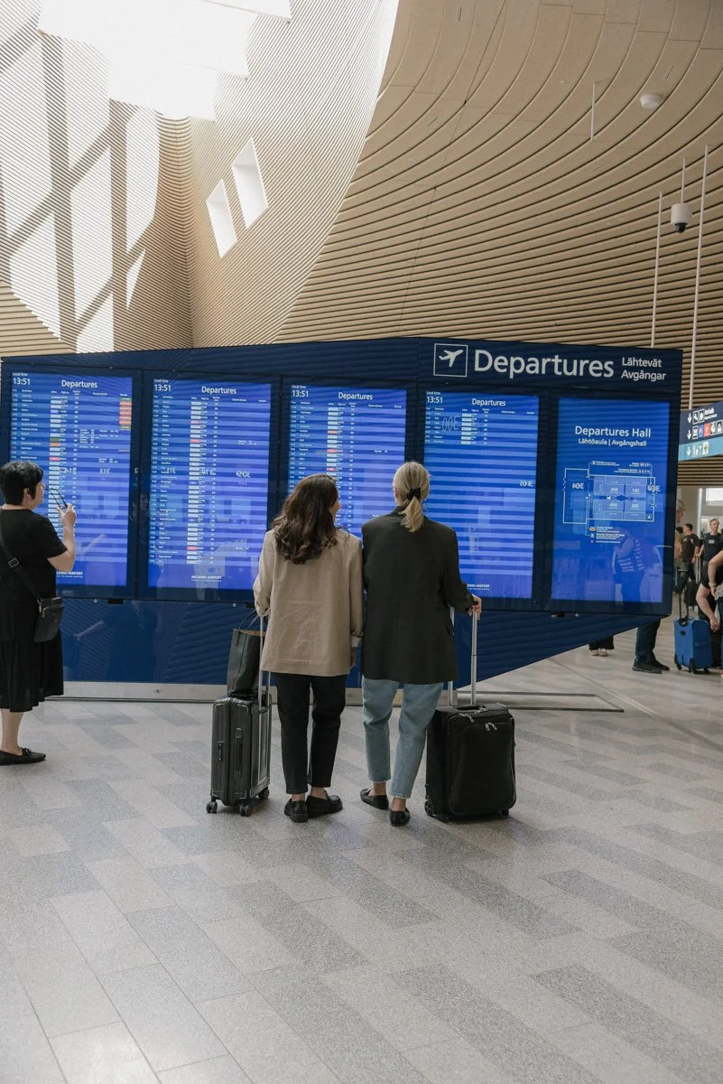 Neam Societyn yrityskuvaus lentokentällä. Kaksi naisyrittäjää seisoo matkalaukkuineen infotaulun edessä.