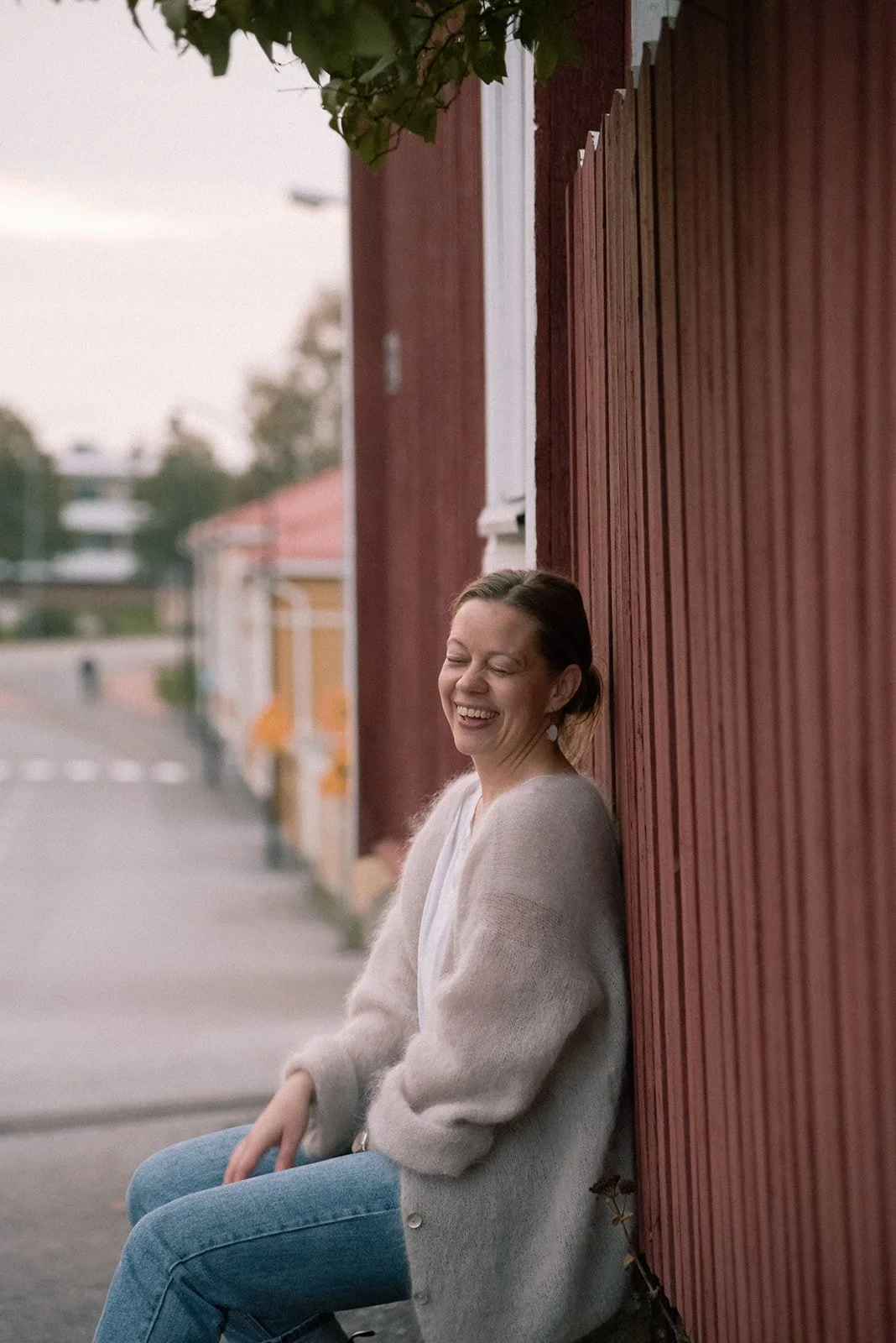 Brändikuva, jossa iloinen naisyrittäjä nojaa punaisen puutalon seinään.