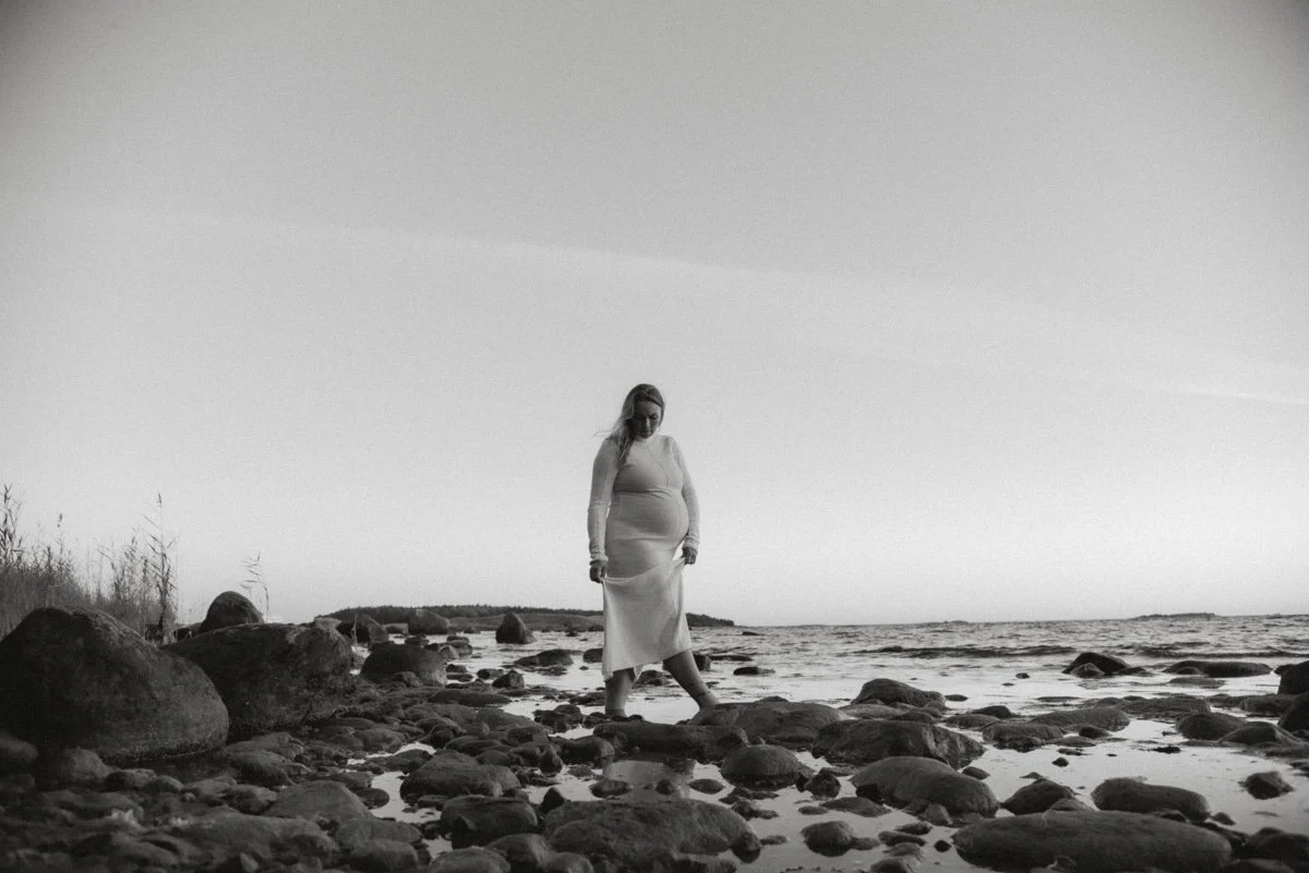 Raskaana oleva nainen kävelee rantakivikossa vaalea mekko yllään. Kuvannut Heidi Väisänen.