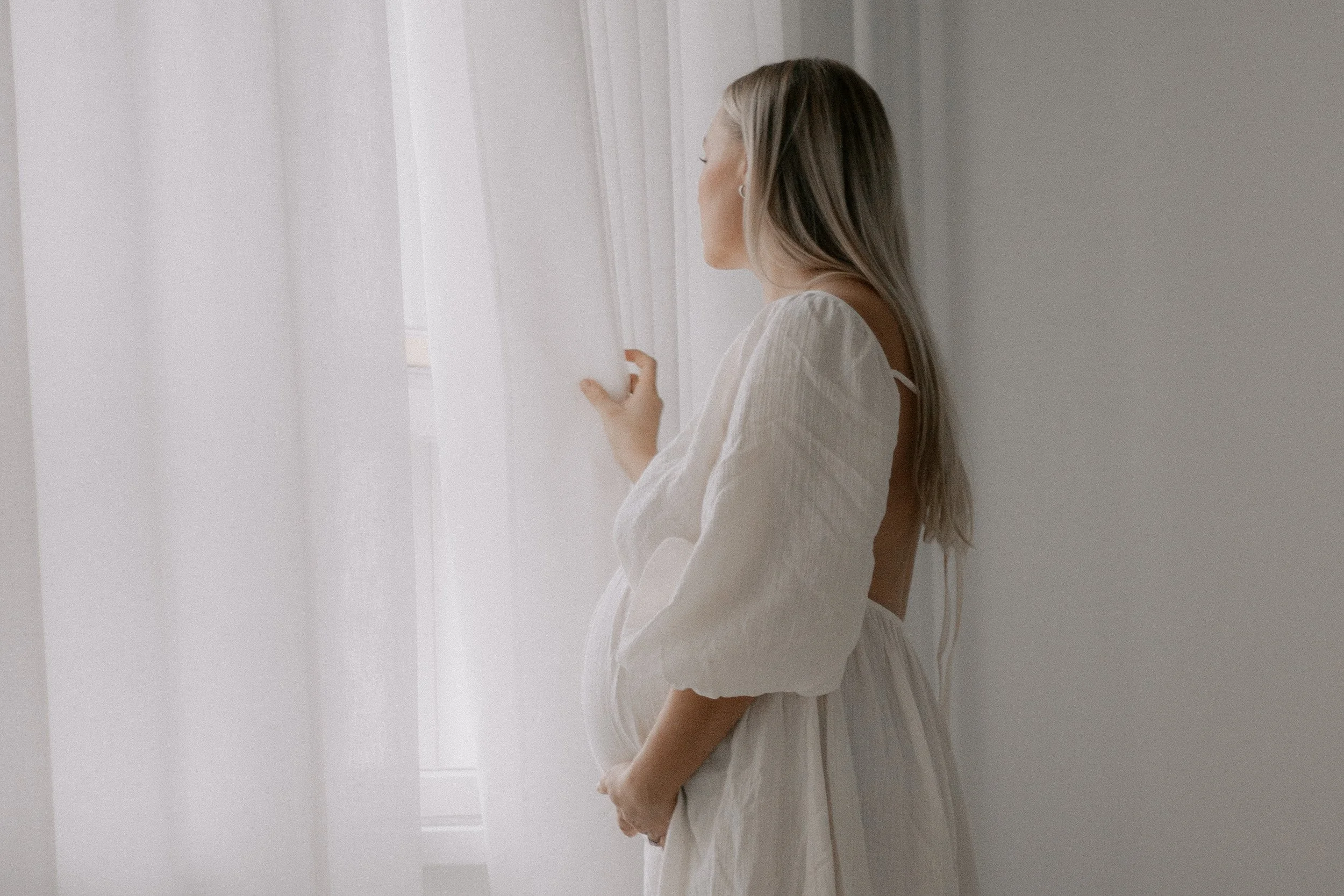 Raskaana oleva äiti kurkistaa ulos ikkunasta ja raottaa vaaleita verhoja. Odotusajankuvaus Heidi Väisäsen studiolla Keravan Saviossa.