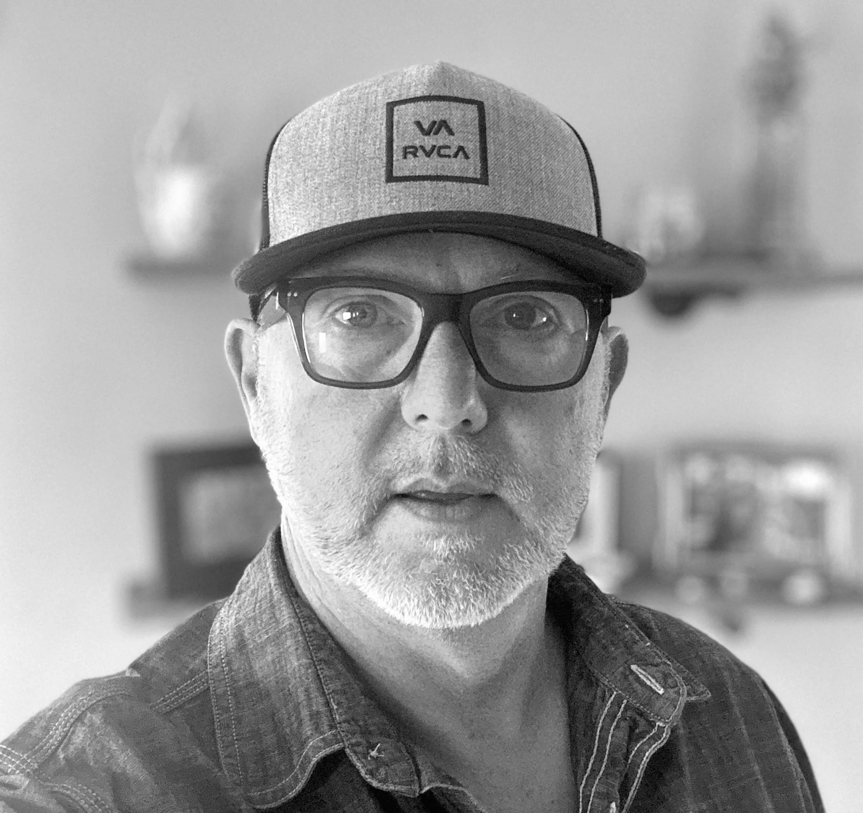 A black and white photo of a man with glasses and a beard, wearing a cap with a logo and a denim jacket.