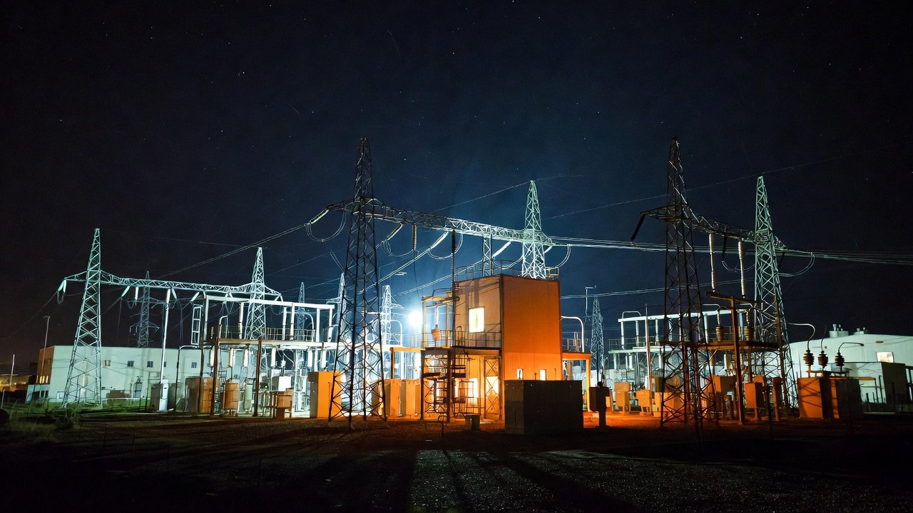 Electric power substation at night with illuminated structures and power lines against the dark sky.