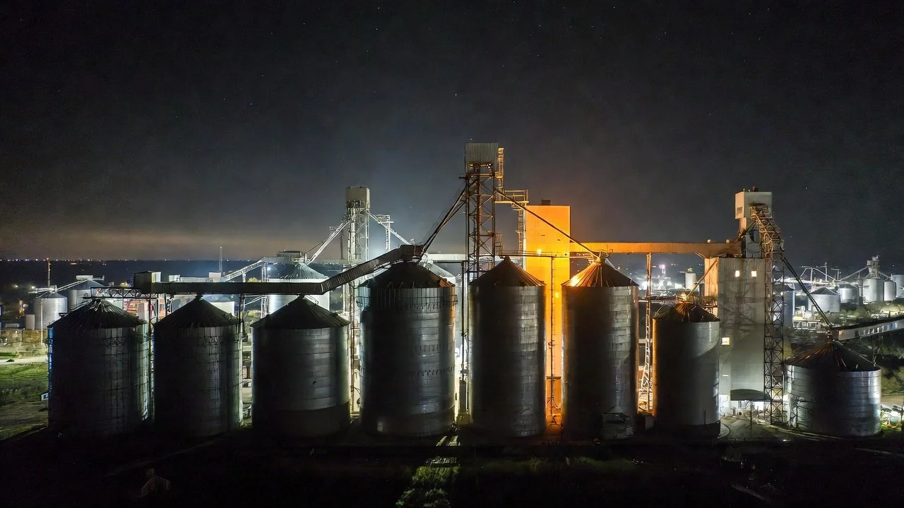 Night view of an industrial grain storage facility with silos and conveyor structures illuminated by artificial lighting