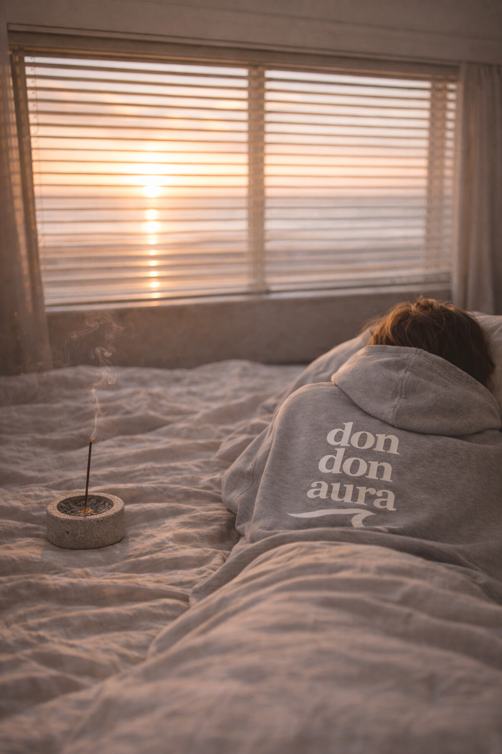Person lying on bed in front of window with blinds, sunrise or sunset lighting, smoking incense in a small holder, wearing a hoodie with 'don don aura' printed on the back.
