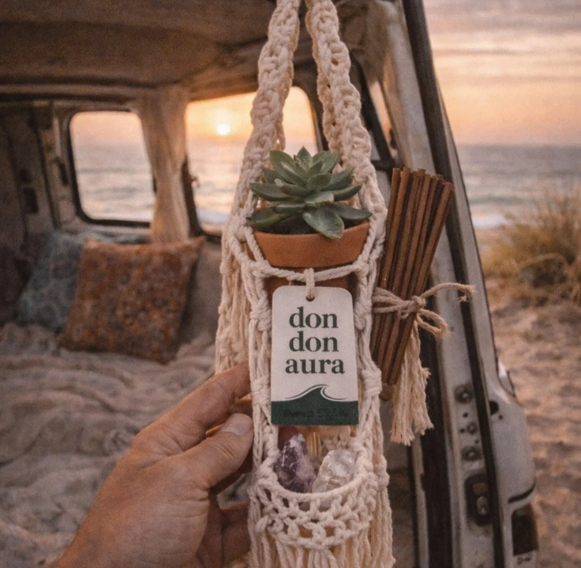A person holding a macrame plant holder with a succulent, a tag reading 'don don aura', and crystals, with a sunset view of the ocean in the background inside a camper van.