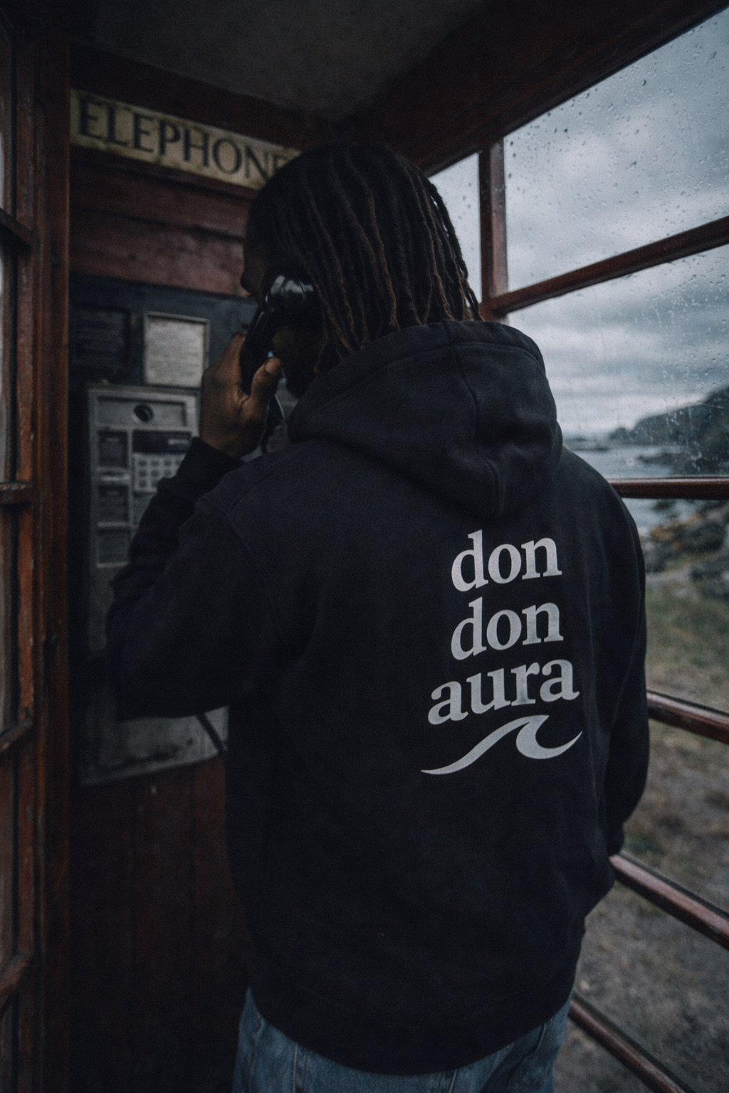 A person with dreadlocks wearing a black hoodie with white text that reads 'don don aura' and a wave symbol, standing inside a wooden phone booth during a rainy day, talking on a black telephone.