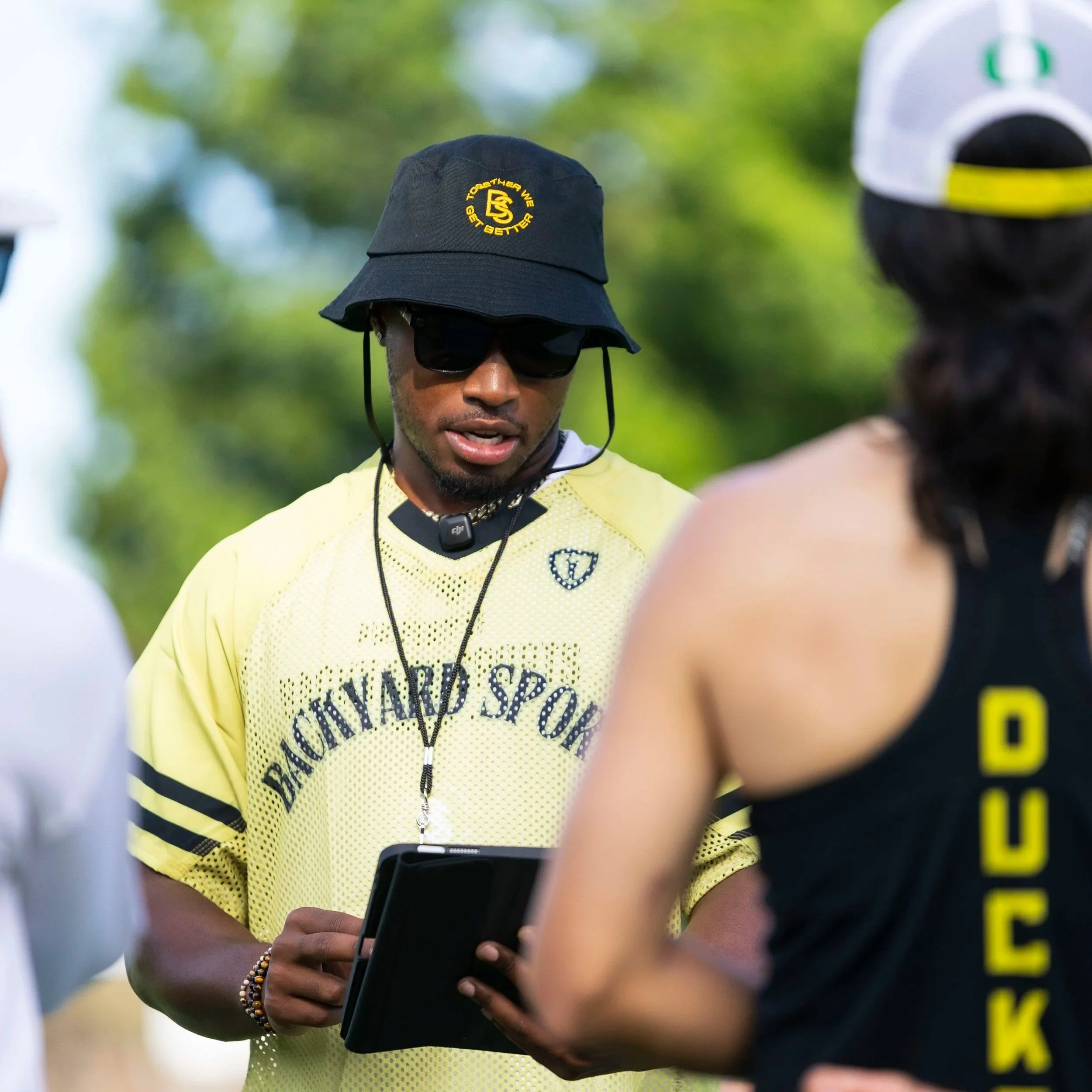 Brian Saulsberry wearing a yellow Backyard Sports Lacrosse t-shirt, black hat with and sunglasses, standing outdoors with a tablet in his hand, talking to a lacrosse players and a College D1 coach wearing a black Oregon Ducks tank top and white cap,