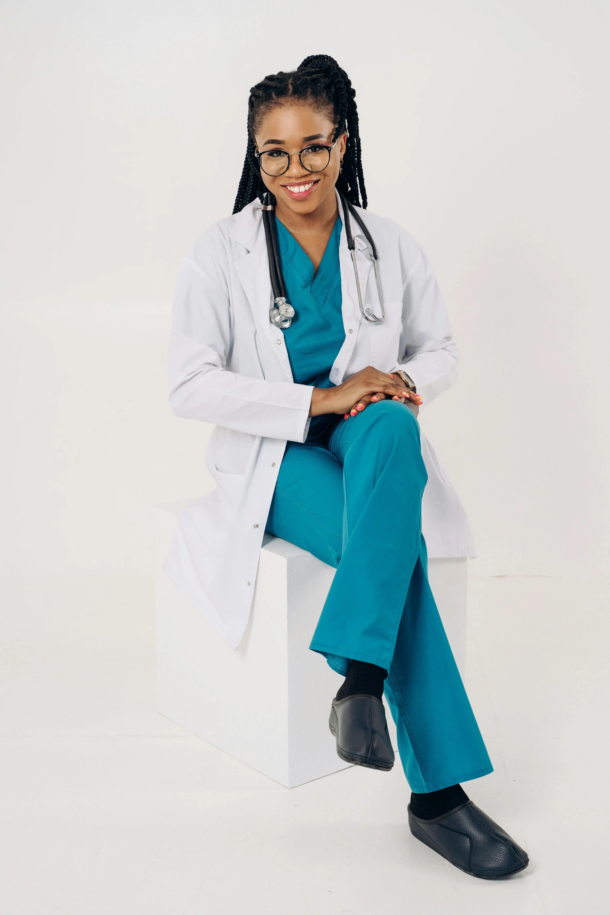 A smiling Black female doctor with glasses, wearing a white coat and teal scrubs, seated on a white block against a plain white background, with a stethoscope around her neck.
