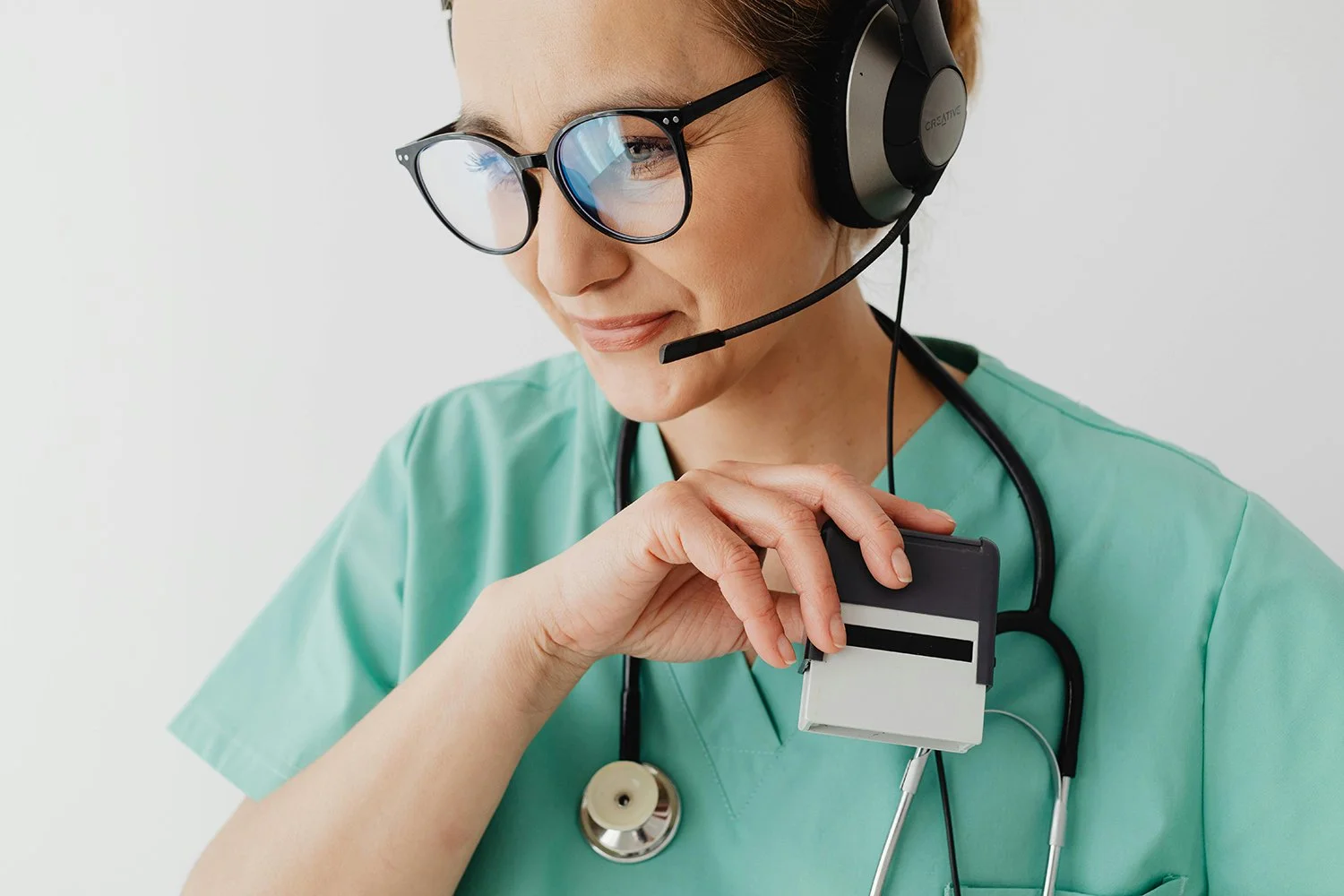 A female healthcare professional wearing glasses, a headset, and a stethoscope around her neck, holding a badge or credential card.