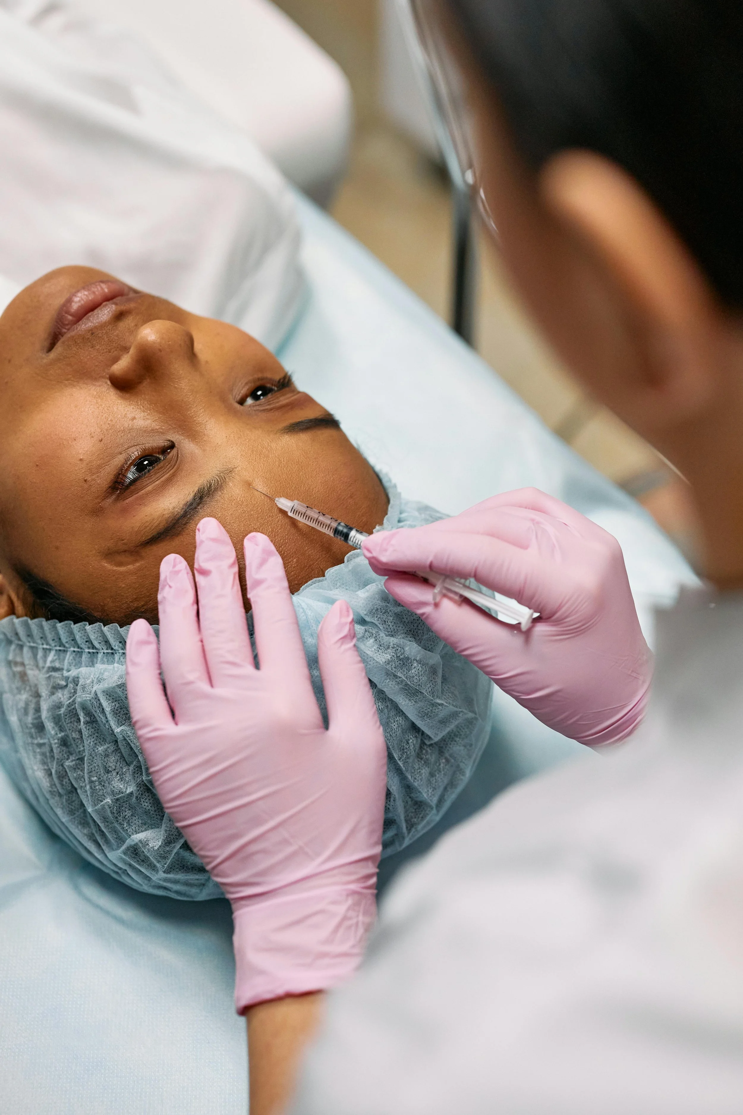 A person in a medical gown and pink gloves receives an injection in their forehead, lying on a hospital bed.
