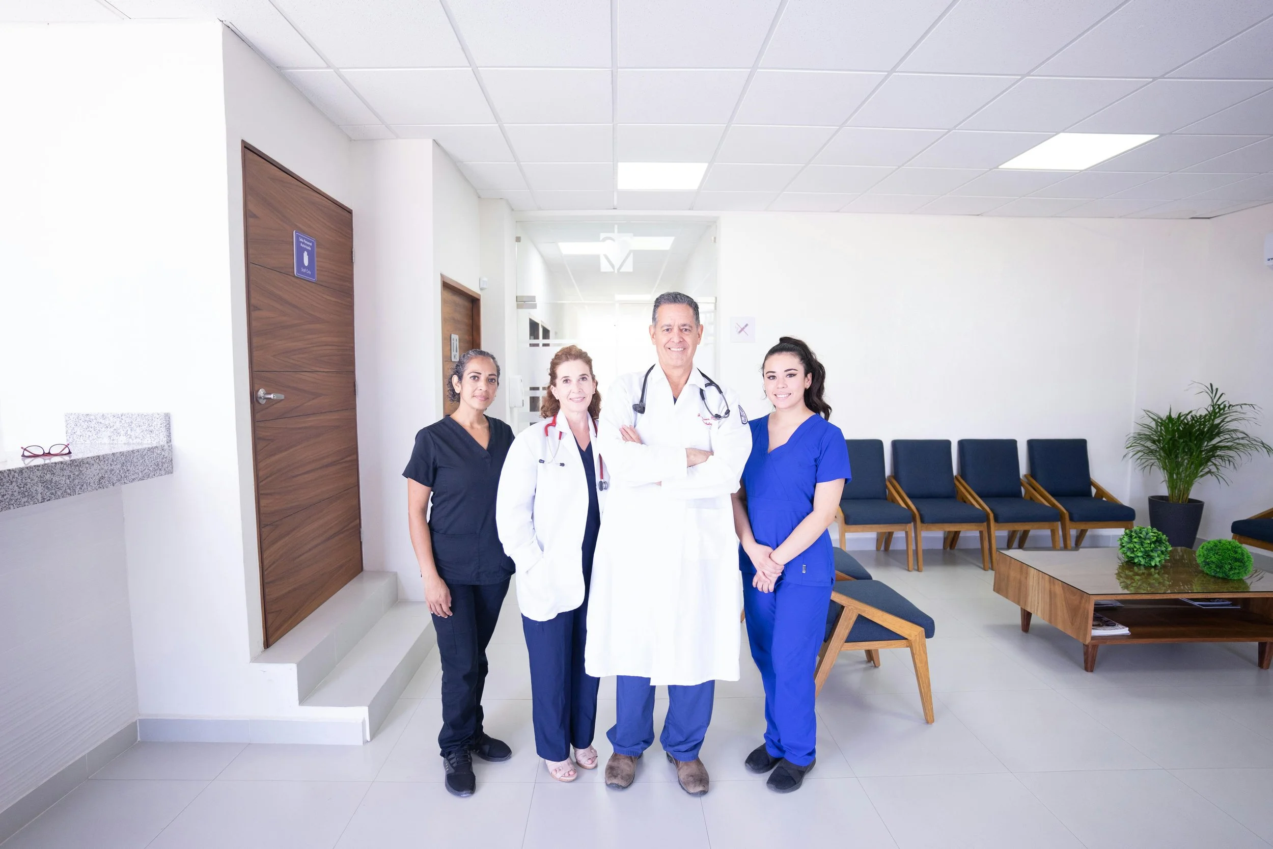Group of four healthcare professionals, including two women in black and blue scrubs, a woman in a white coat, and a man in a white coat with a stethoscope, standing together in a medical facility lobby with chairs and plants in the background.