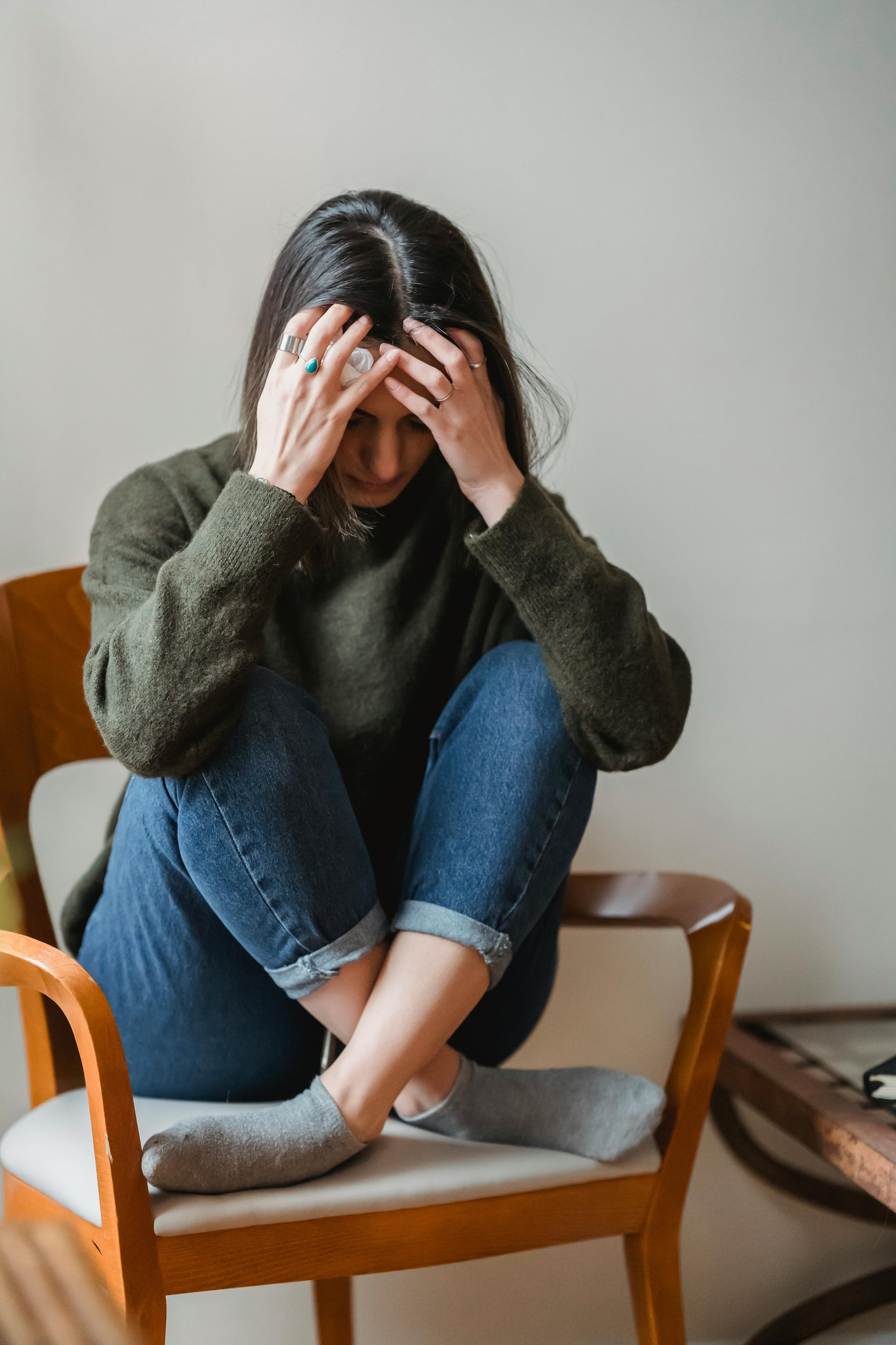 A woman sitting on a chair with her knees up, holding her head with her hands, looking down, appearing distressed.