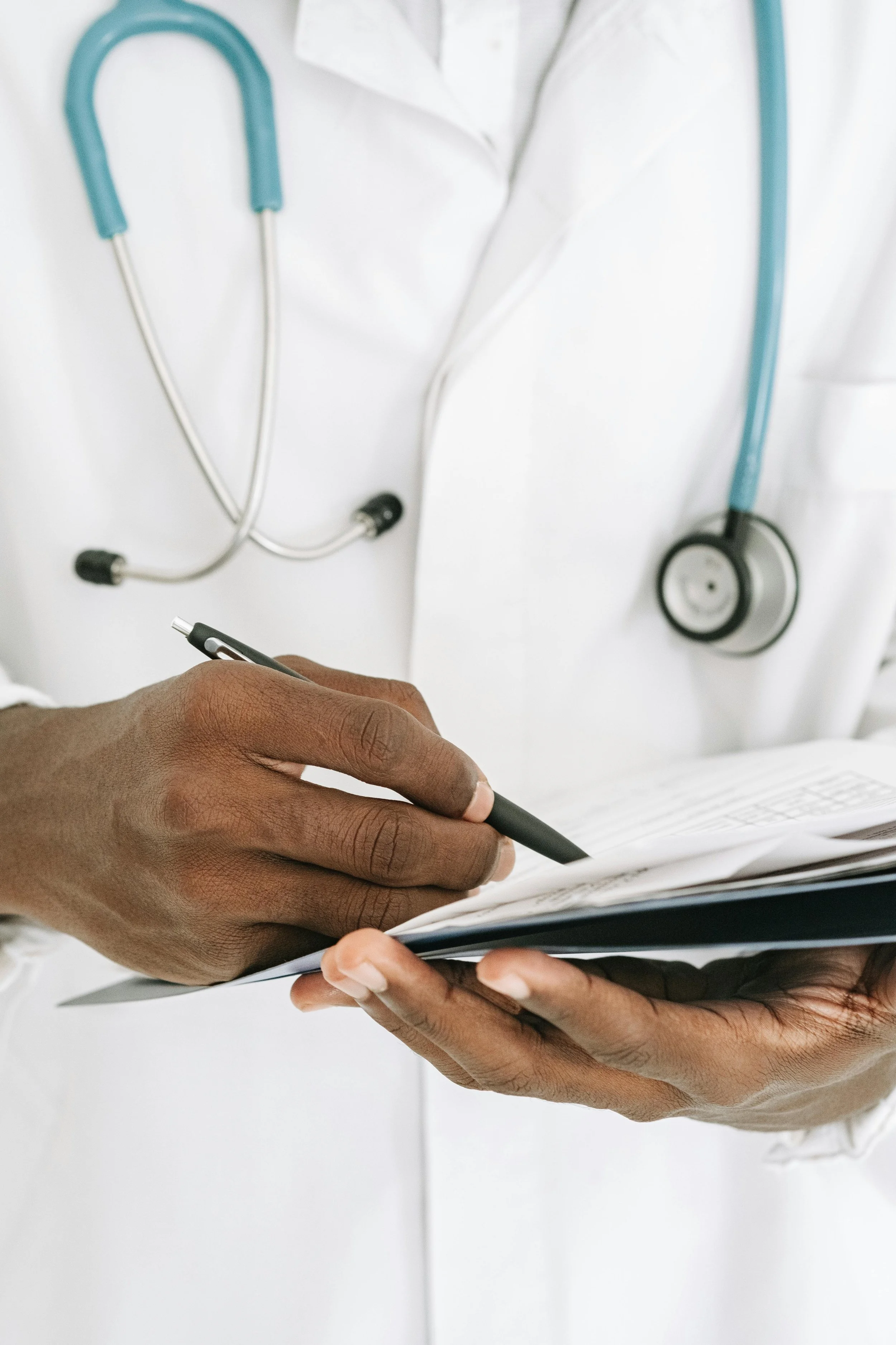 Close-up of a doctor or healthcare professional writing on a medical chart with a pen, wearing a white coat and stethoscope around neck.
