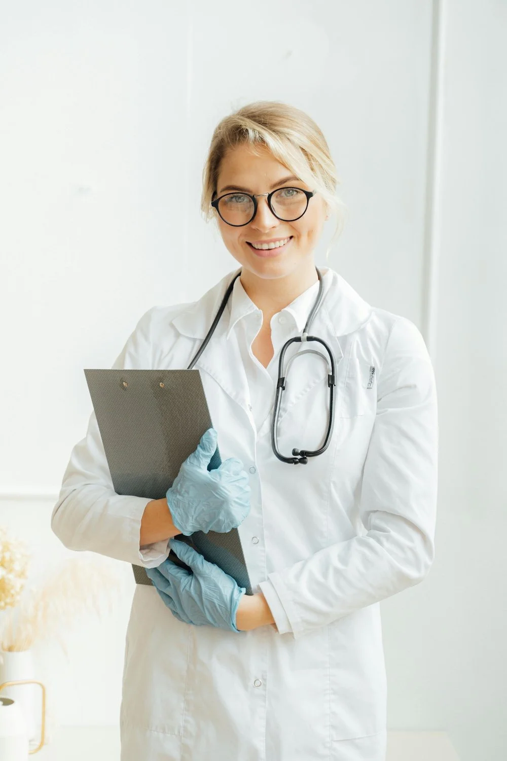 A female doctor wearing glasses, a white lab coat, and a stethoscope around her neck, smiling and holding a clipboard with both hands, wearing blue gloves.