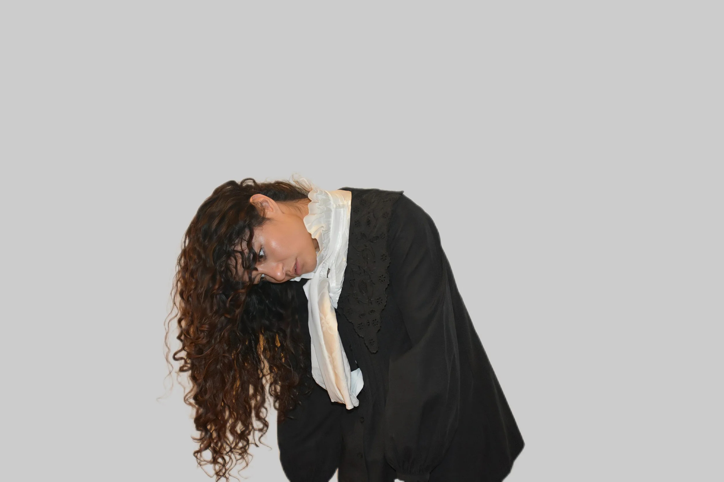 Donna con capelli riccioli scuri, vestita con abito nero e camicia bianca, si piega in avanti contro uno sfondo grigio.