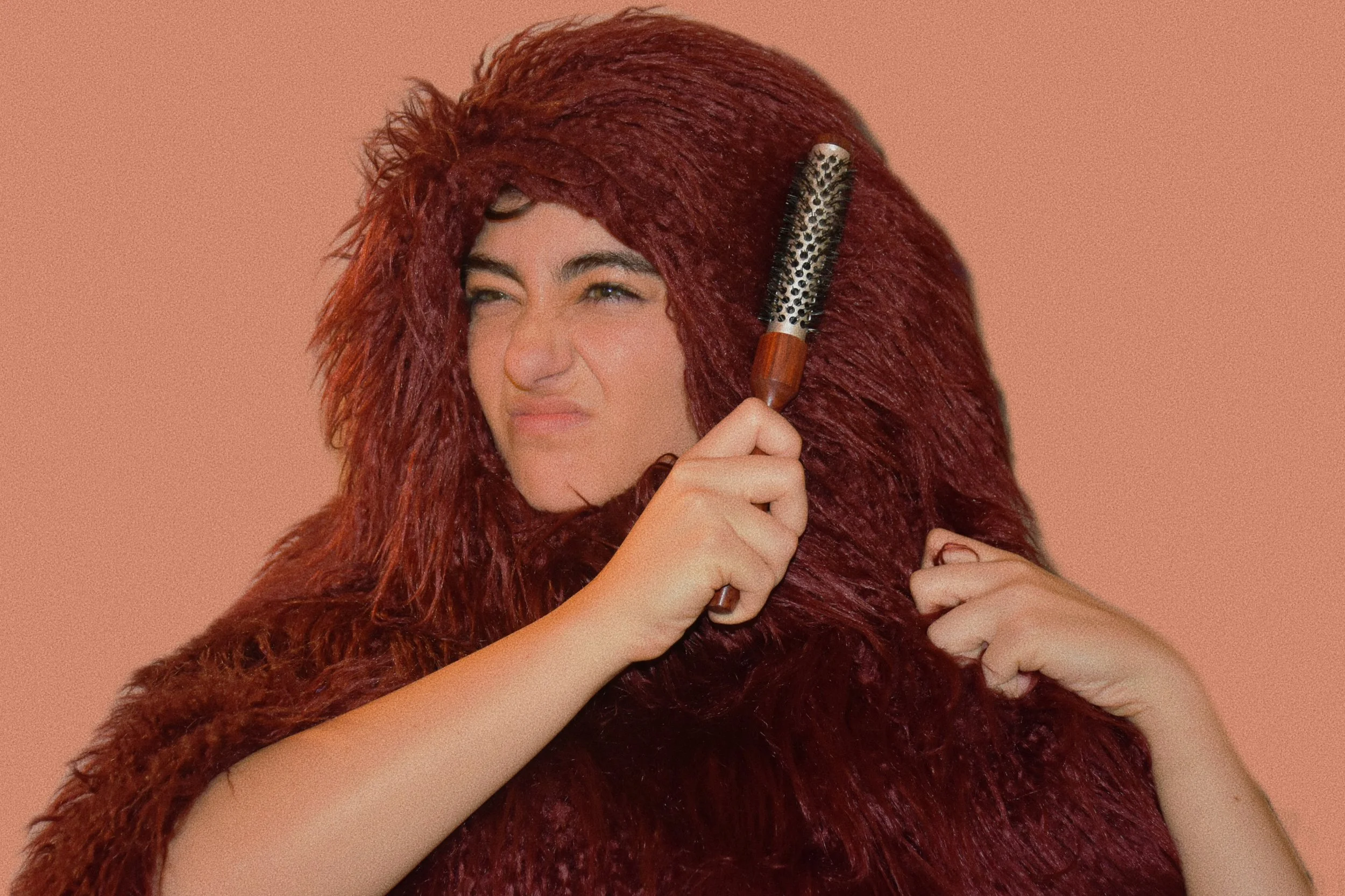 Donna con capelli rossi, avvolta in una coperta dello stesso colore, che si pettina i capelli con una spazzola, guardando in avanti con un'espressione accigliata.