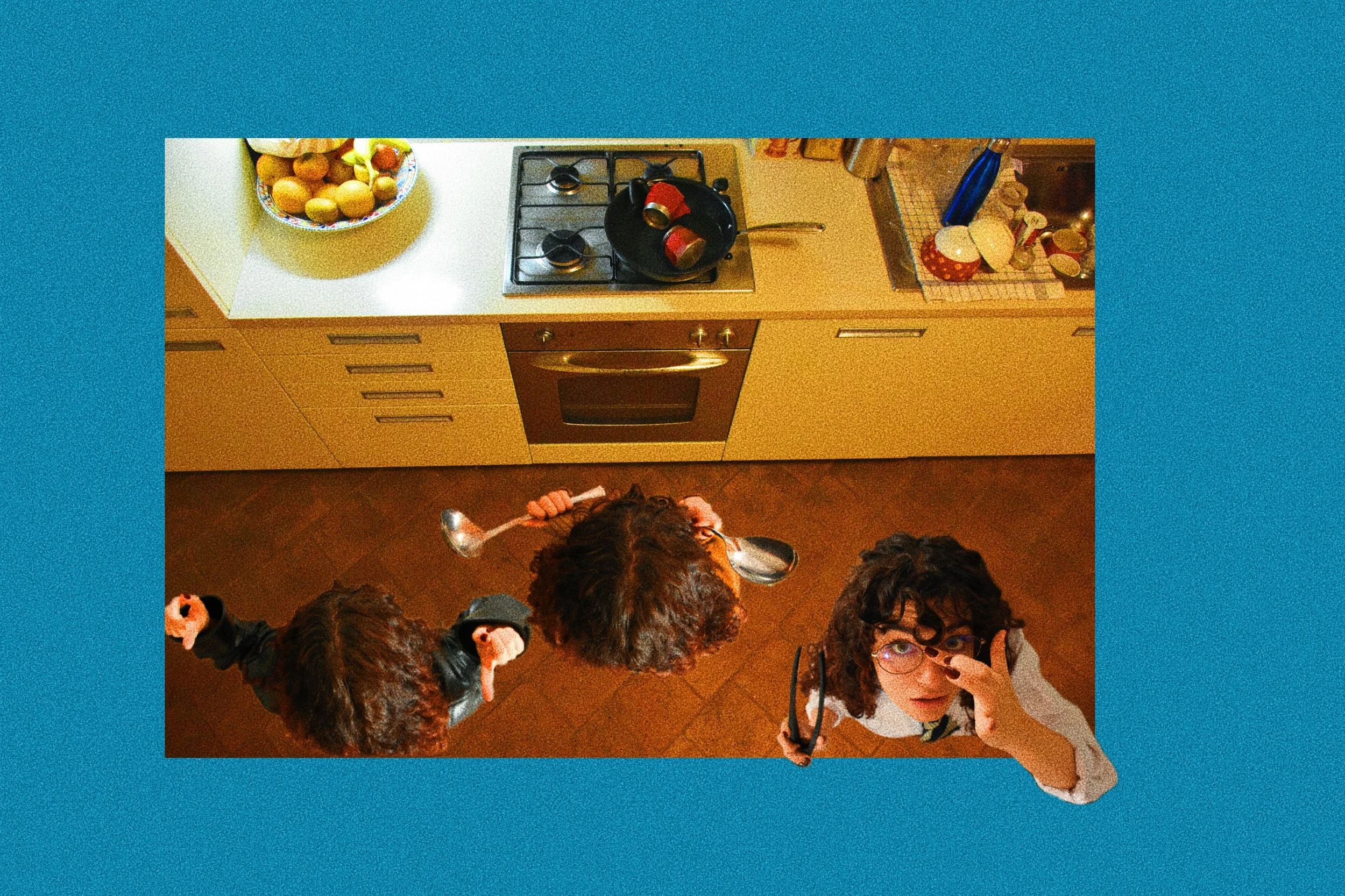 Vista dall'alto di due bambini in cucina, uno con occhiali e un'altra che si pulisce gli occhiali, con cucina e piano di lavoro visibili.