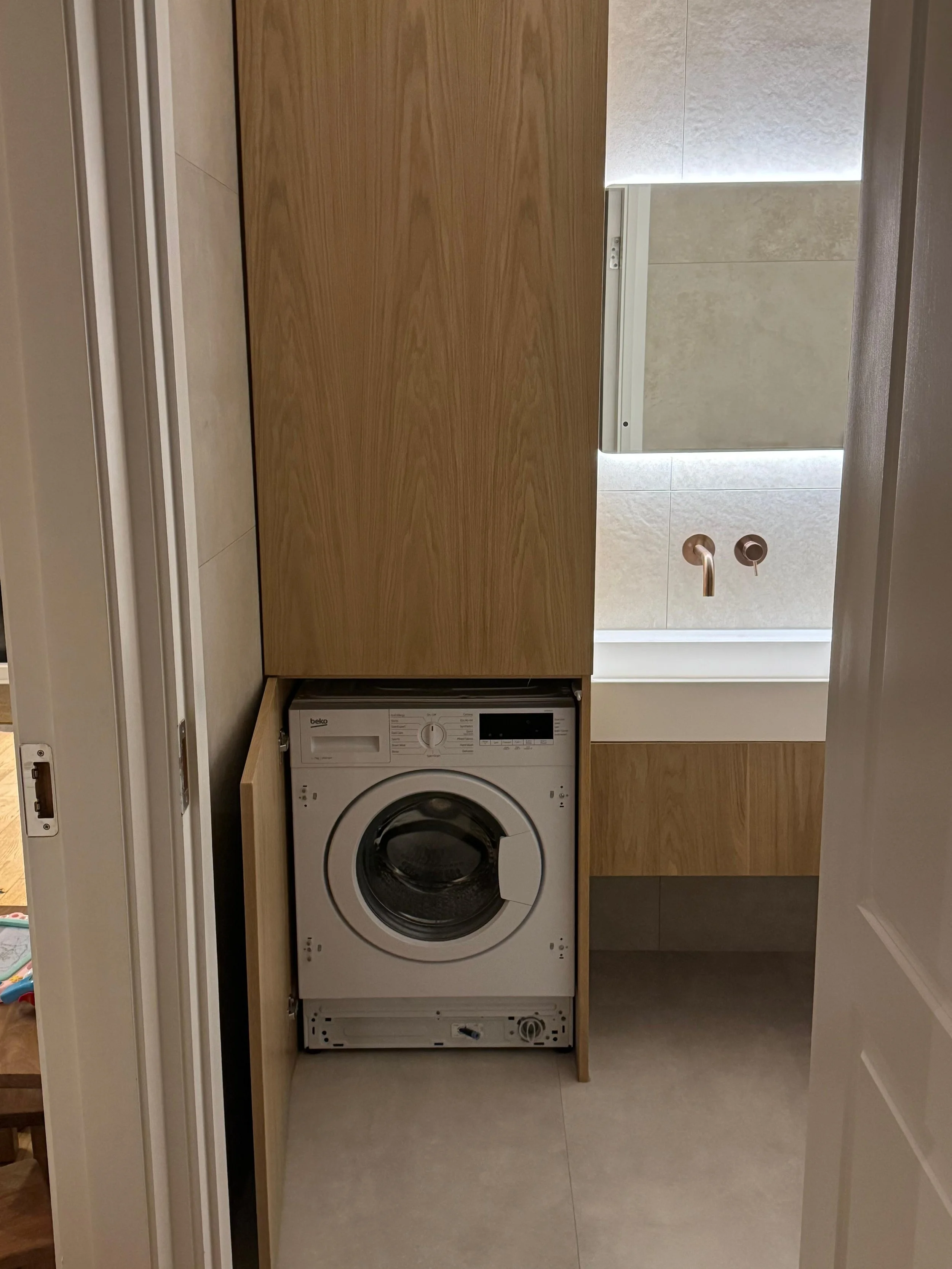 A front-loading washing machine installed inside a wooden cabinet in a bathroom or laundry room, with beige tile flooring and a bathroom vanity with a mirror and faucet in the background.