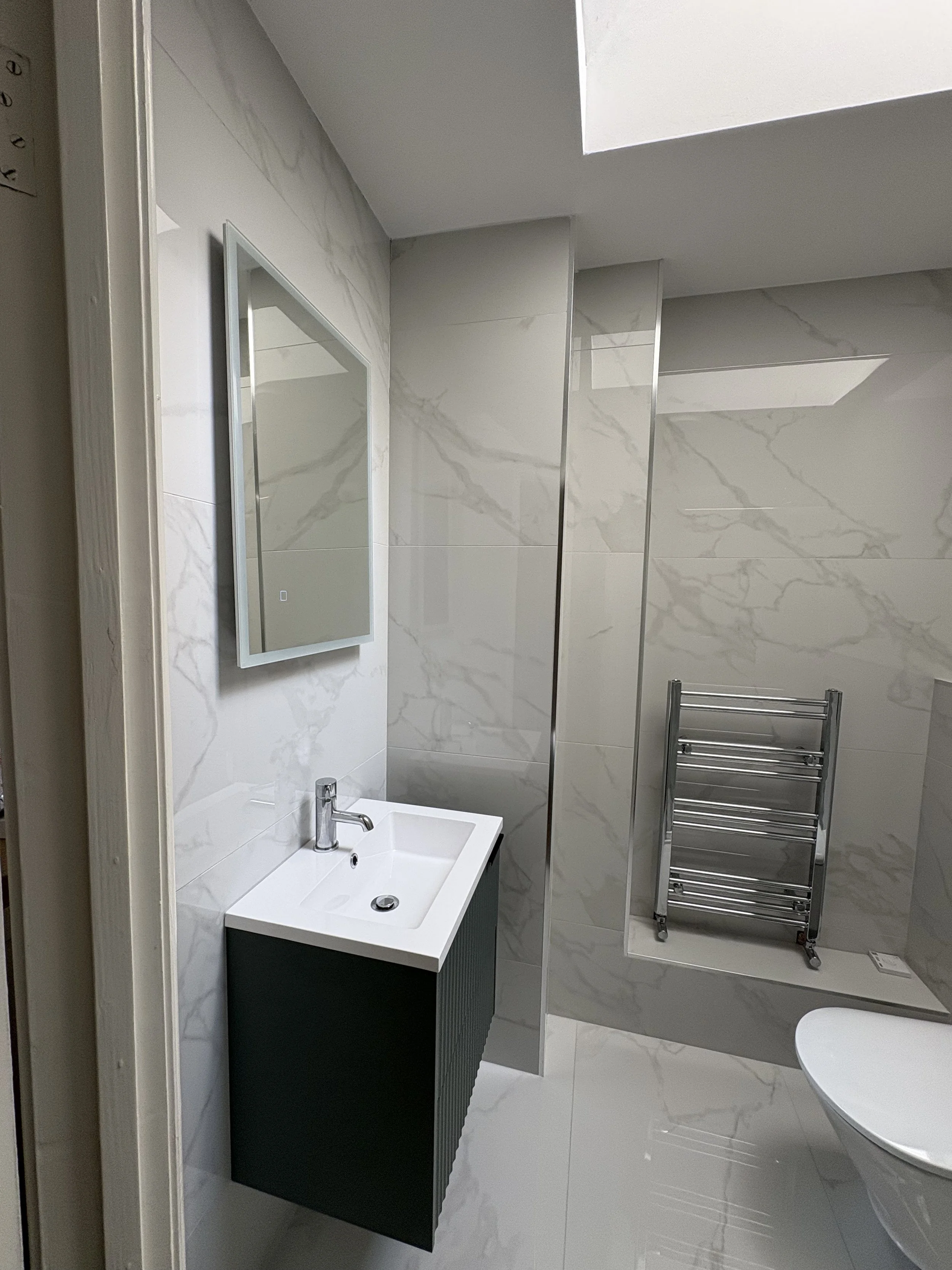 Bathroom with white marble walls, a small white sink with a black cabinet, a rectangular mirror, a metal towel rack, and a white toilet.