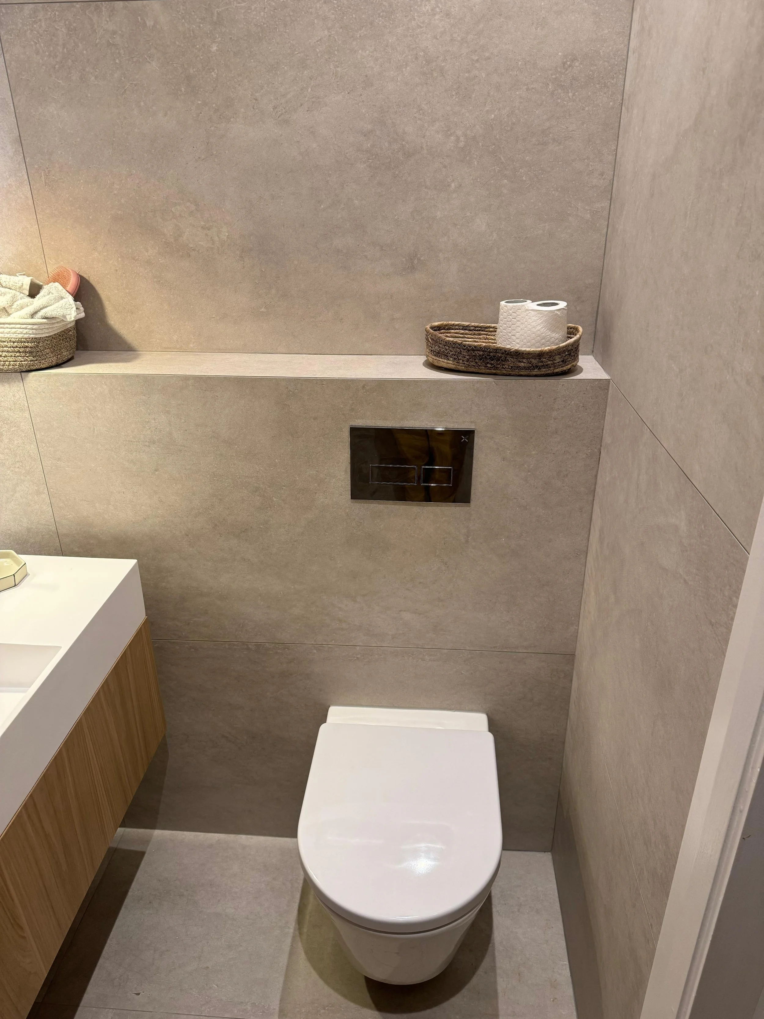 A modern bathroom with beige tiled walls and floor, a white toilet, a wooden cabinet with a white countertop to the left, and a shelf holding a basket with towels and a tray with toilet paper rolls.