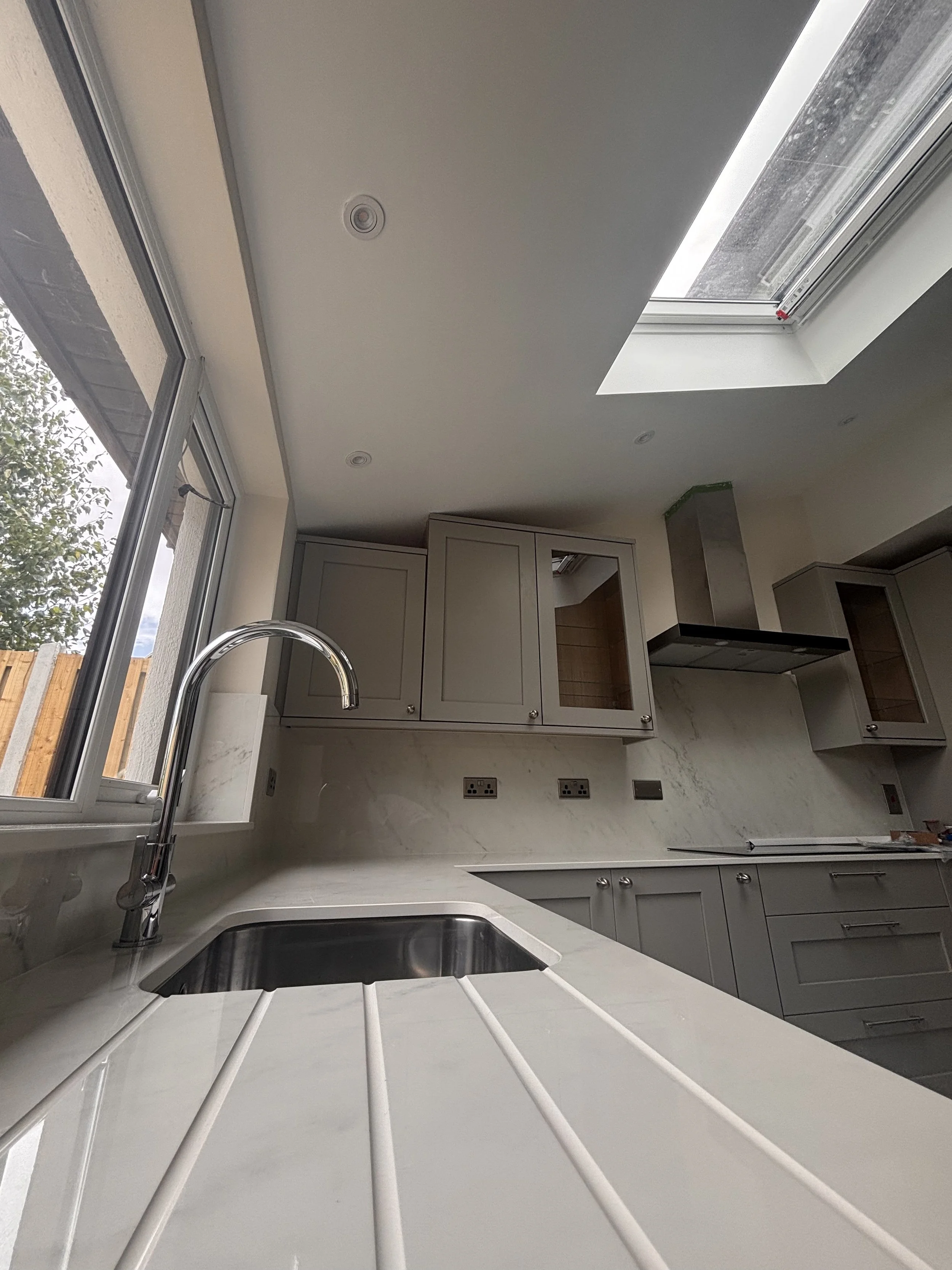 Modern kitchen with white countertops, gray cabinets, a stainless steel sink with a chrome faucet, and large windows and skylight allowing natural light.