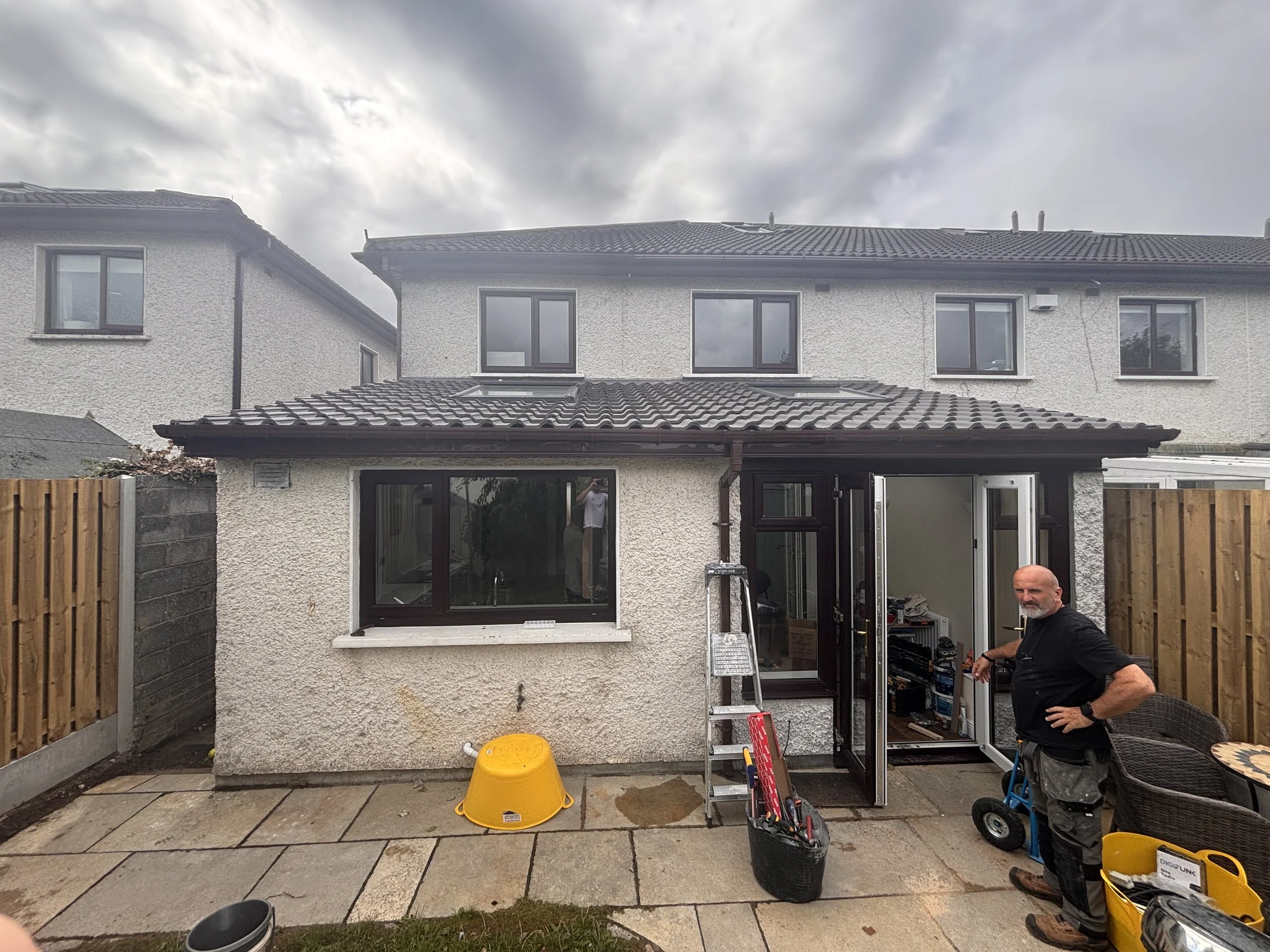 Backyard view of a partly cloudy day showing a two-story house with white textured exterior walls, black window frames, and a tiled roof. There is a man in work clothes standing outside, near an open door, with various tools and equipment nearby.