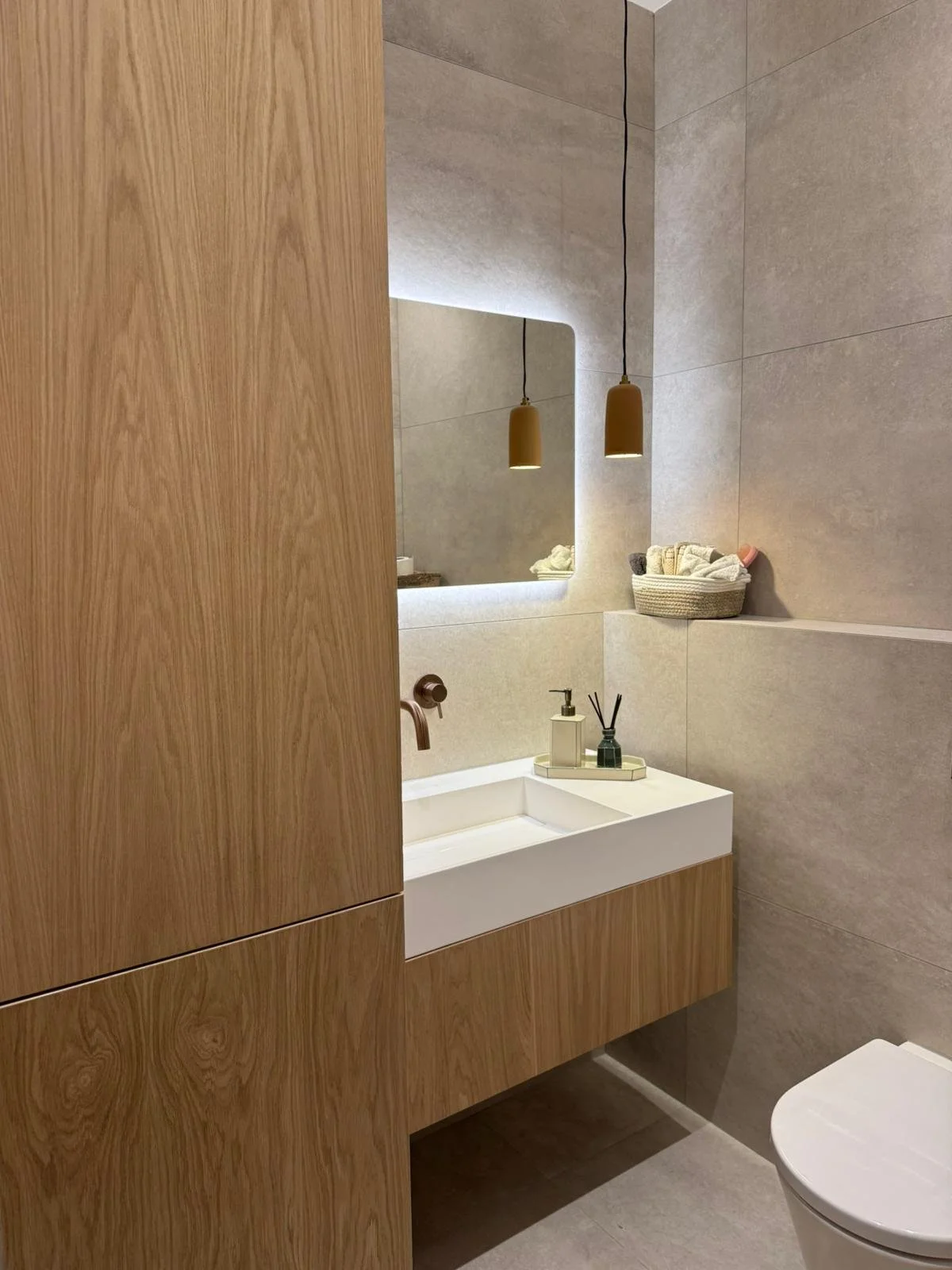 A modern bathroom with a wooden vanity, a white sink, a large rectangular mirror with LED backlighting, beige tiles on the walls, and two hanging pendant lights. A basket with towels and toiletries is on a ledge near the mirror.