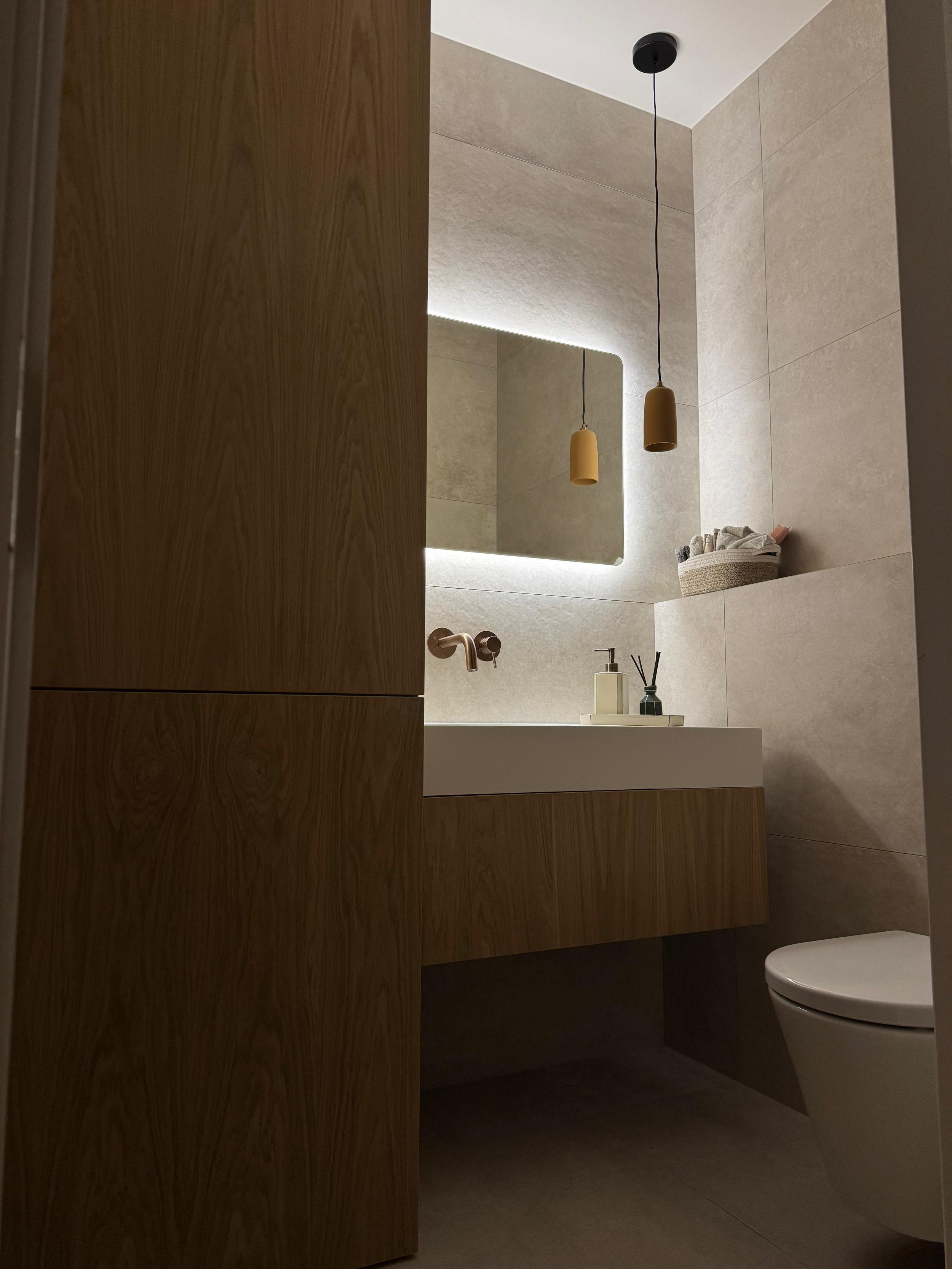 Modern bathroom with beige tiled walls, a large mirror with backlighting, two hanging pendant lights, a white ceramic sink with wooden cabinetry, and a white toilet.