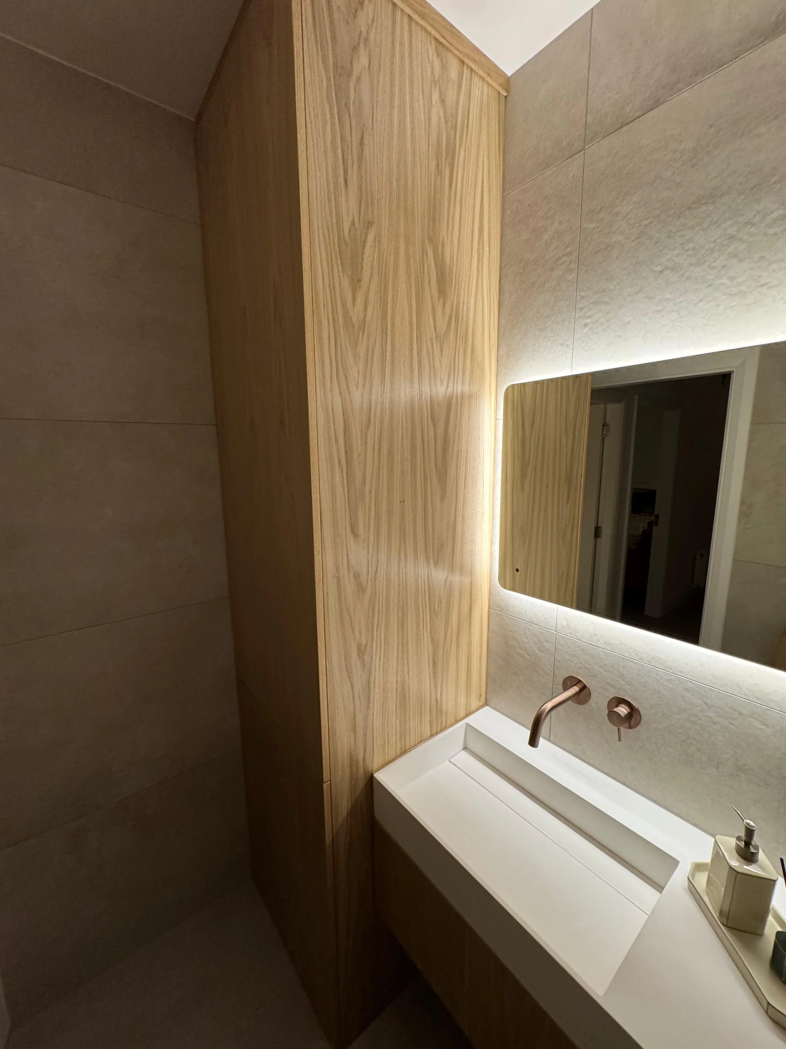 Modern bathroom with a wooden cabinet, white sink, wall-mounted faucet, and a mirror with backlit lighting.