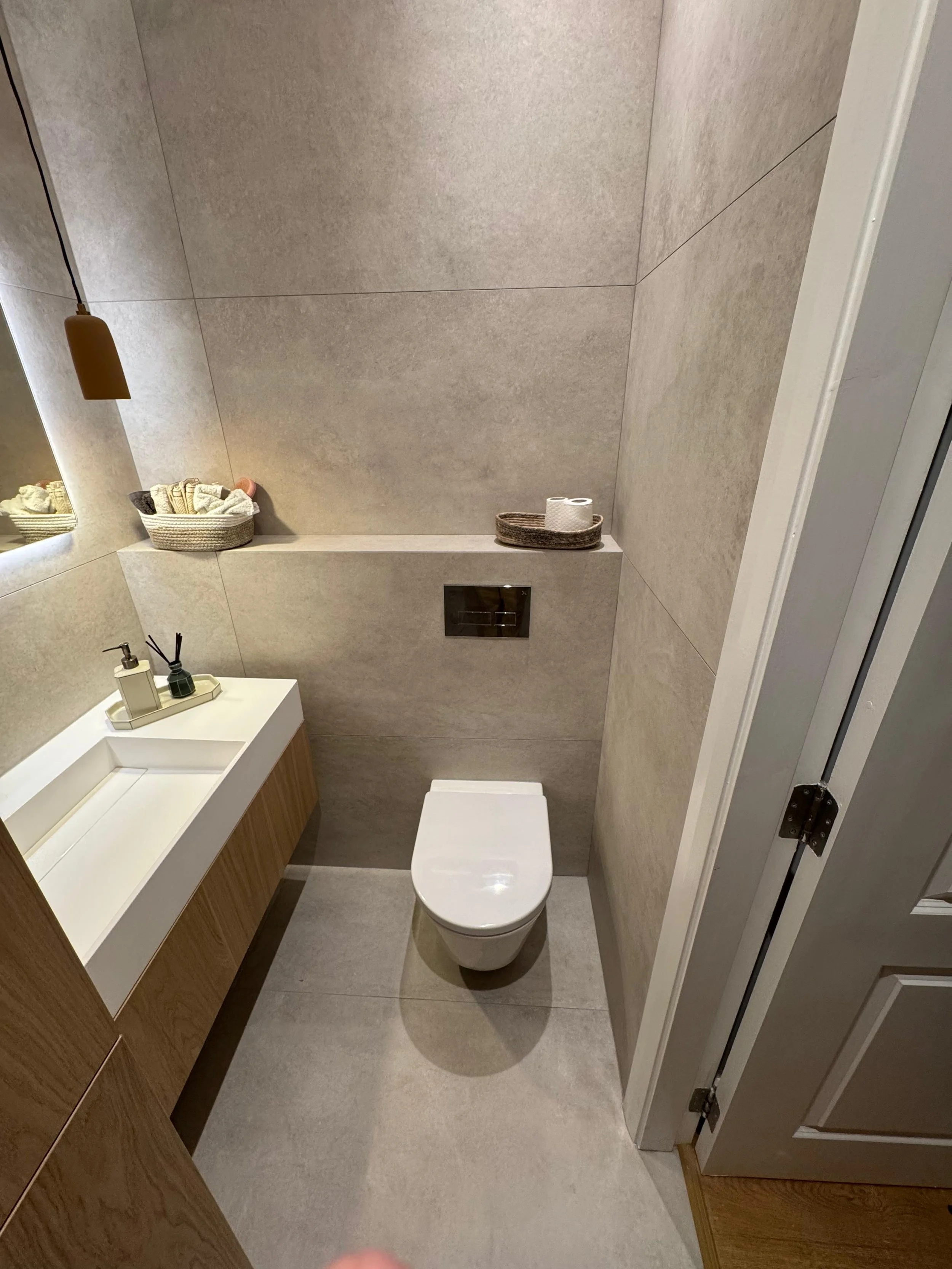 Modern beige bathroom with a wall-mounted toilet, a wooden vanity with a white sink, and minimal decor including baskets and a tray with rolls of toilet paper.