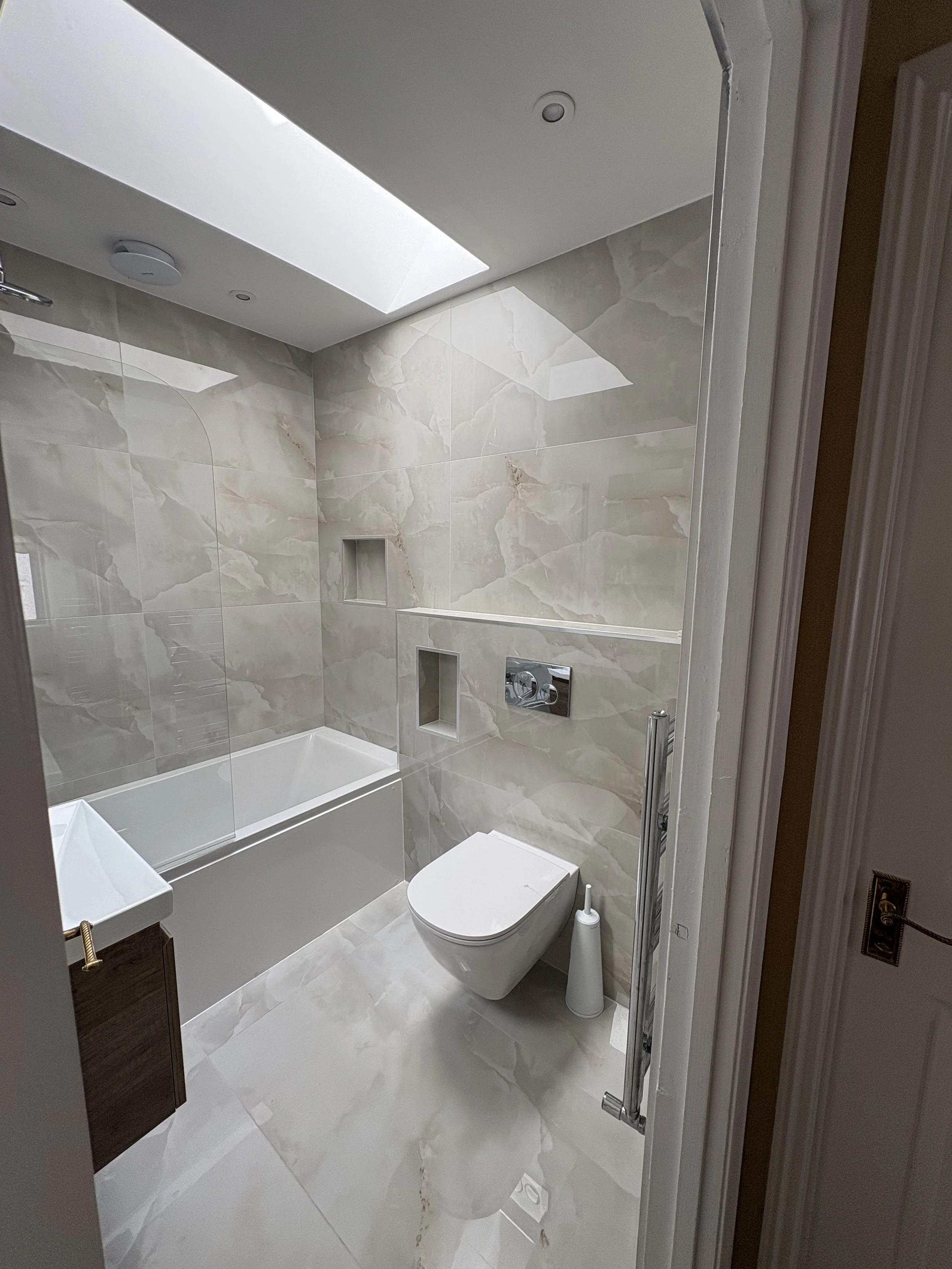 Modern bathroom with beige marble tiles, a built-in bathtub, a wall-mounted toilet, a small sink, a frosted glass window, and skylight.