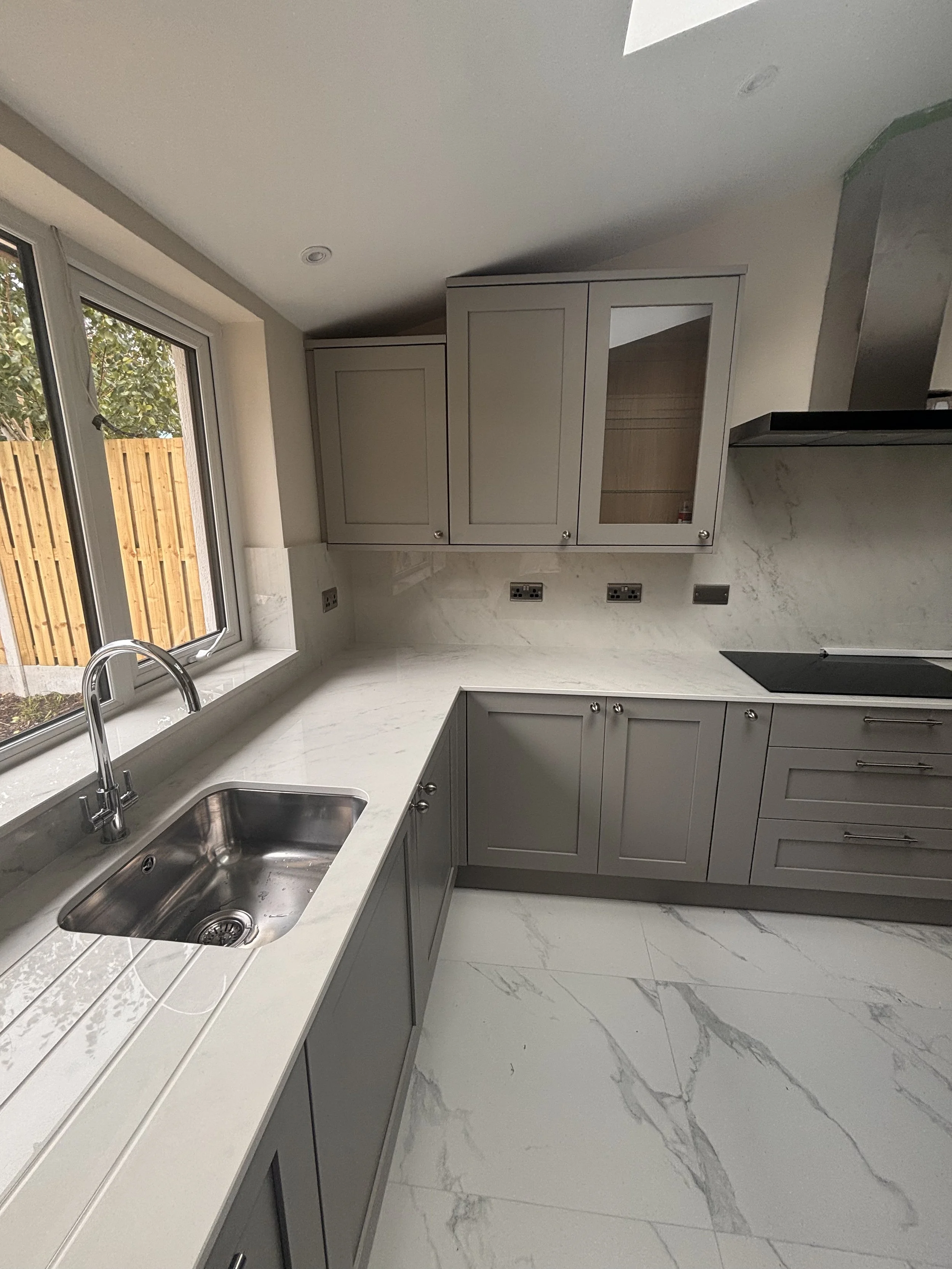 Modern kitchen with gray cabinets, a white marble countertop, a stainless steel sink near a window, and a marble backsplash.