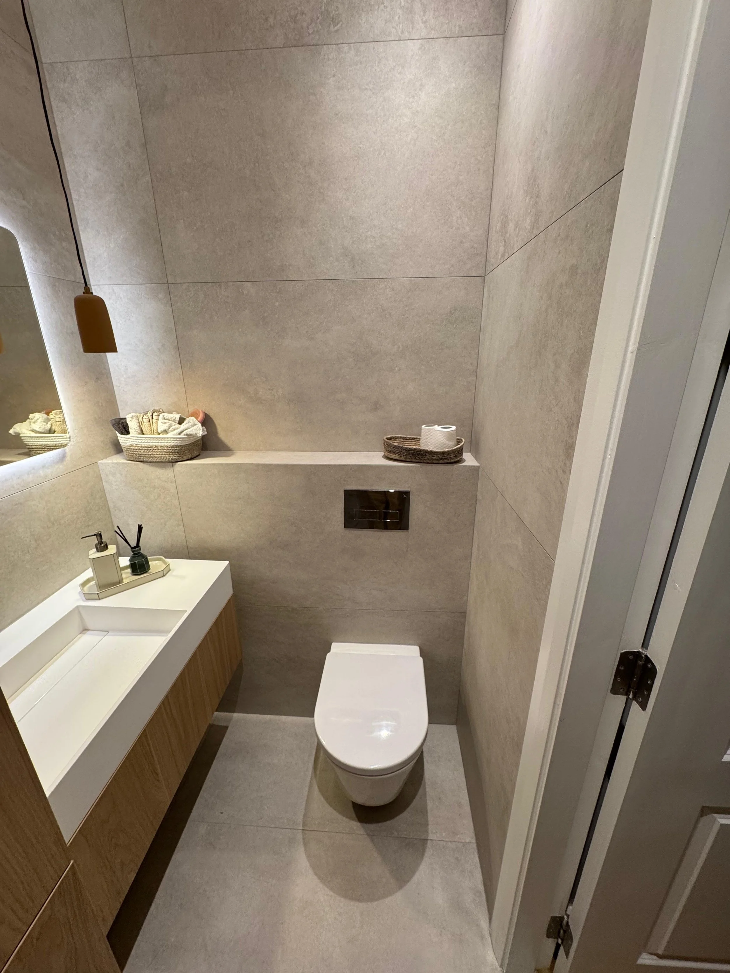 A modern bathroom with a white toilet, beige tiled walls, a wooden vanity with a built-in sink, a mirror, and minimalist decor including a basket of towels and a tray with toiletries, illuminated by a hanging pendant light.