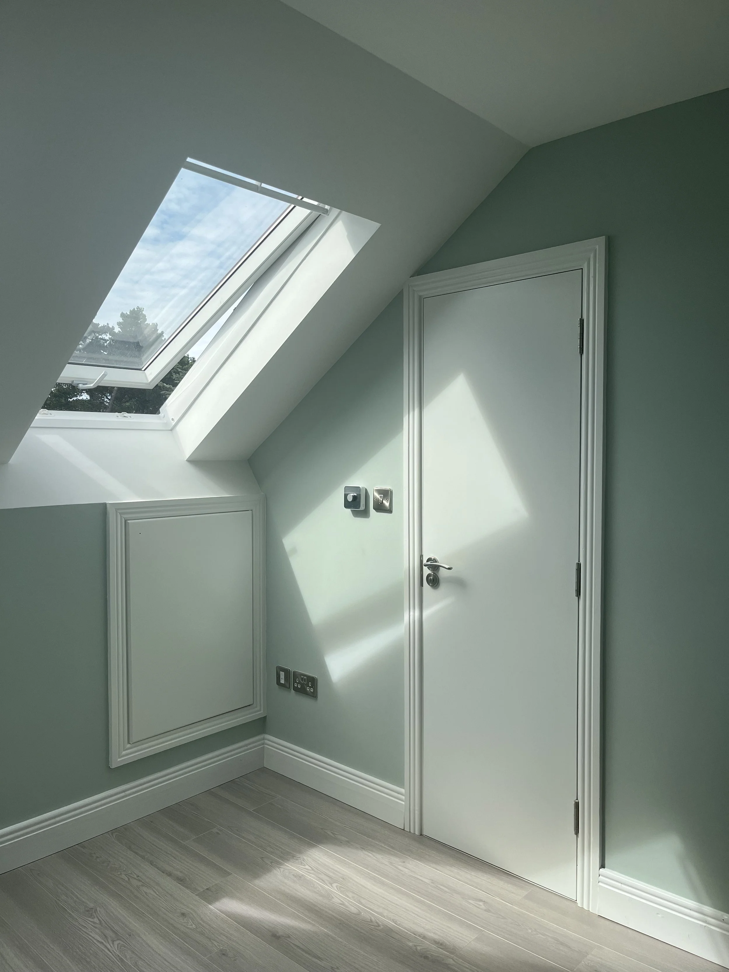 A bright room with a slanted ceiling, a skylight window letting in natural light, a closed white door, and light-colored wooden flooring.