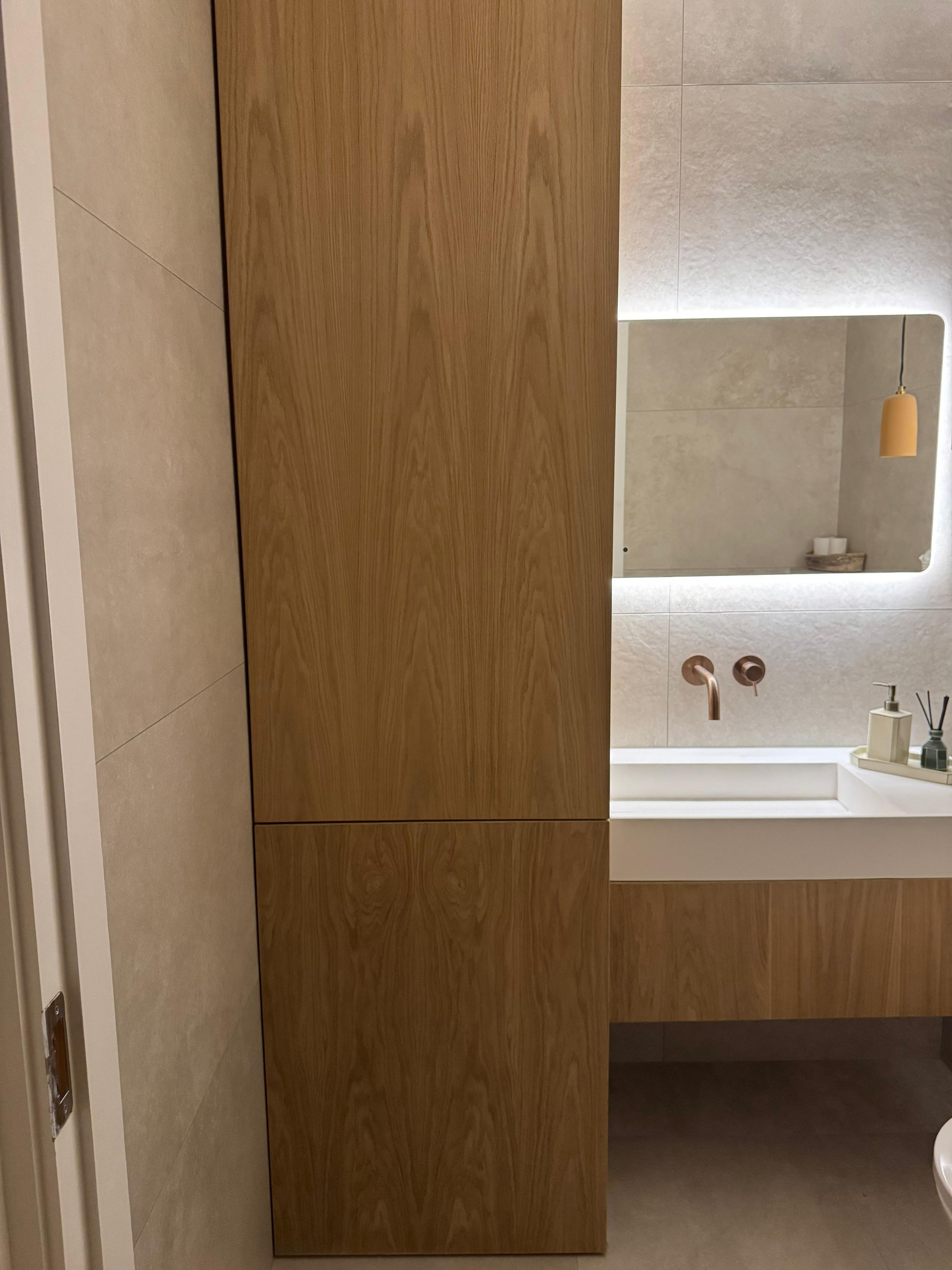 Modern bathroom with a large mirror, white sink, copper faucet, and wooden cabinetry.