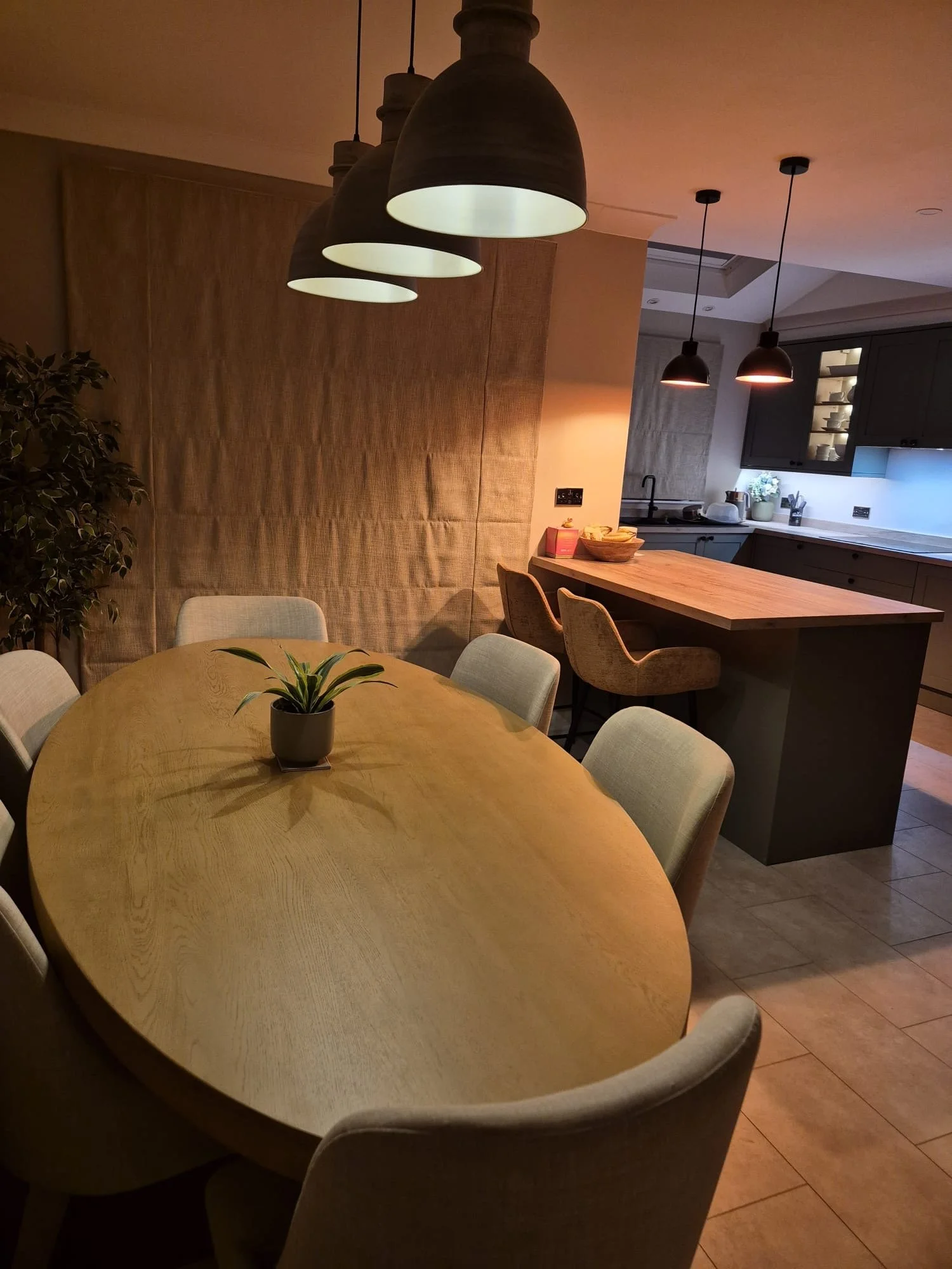 Dinner room with a large wooden oval table surrounded by six light-colored chairs and a small potted plant in the center. Overhead pendant lights hang above the table, and the kitchen area with bar stools, cabinets, and a counter is visible in the background.