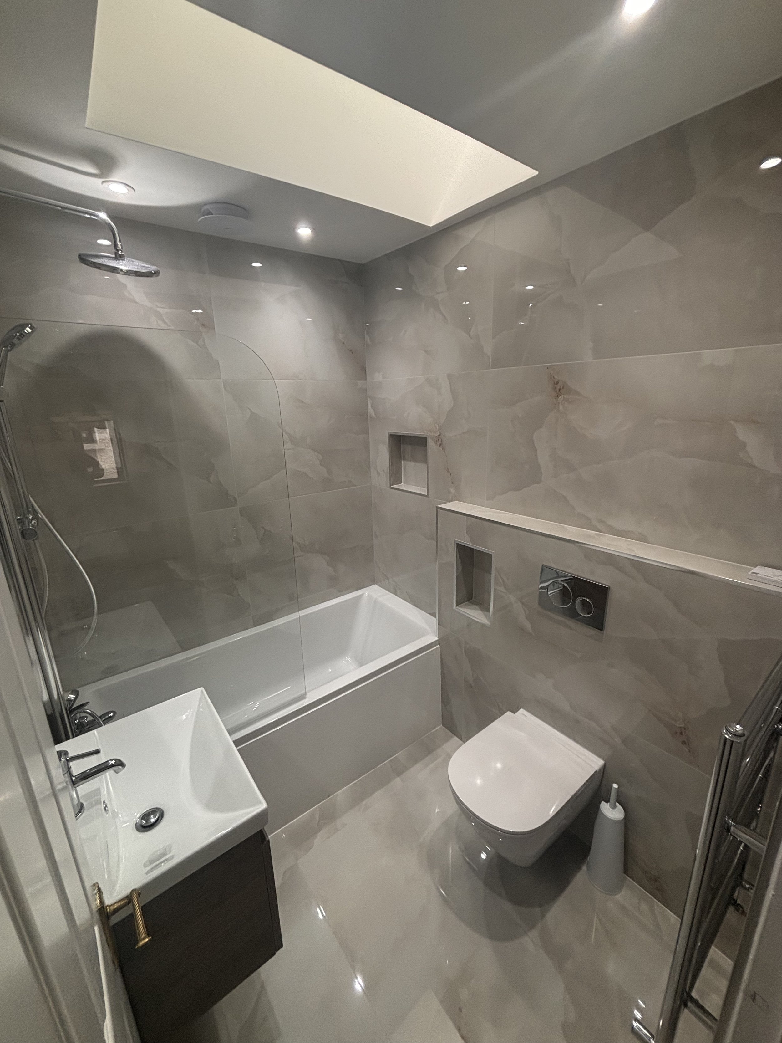 Modern small bathroom with beige marble walls and floors, a white bathtub with a glass partition, a small sink with a wooden cabinet, a wall-mounted toilet, and ceiling lights.