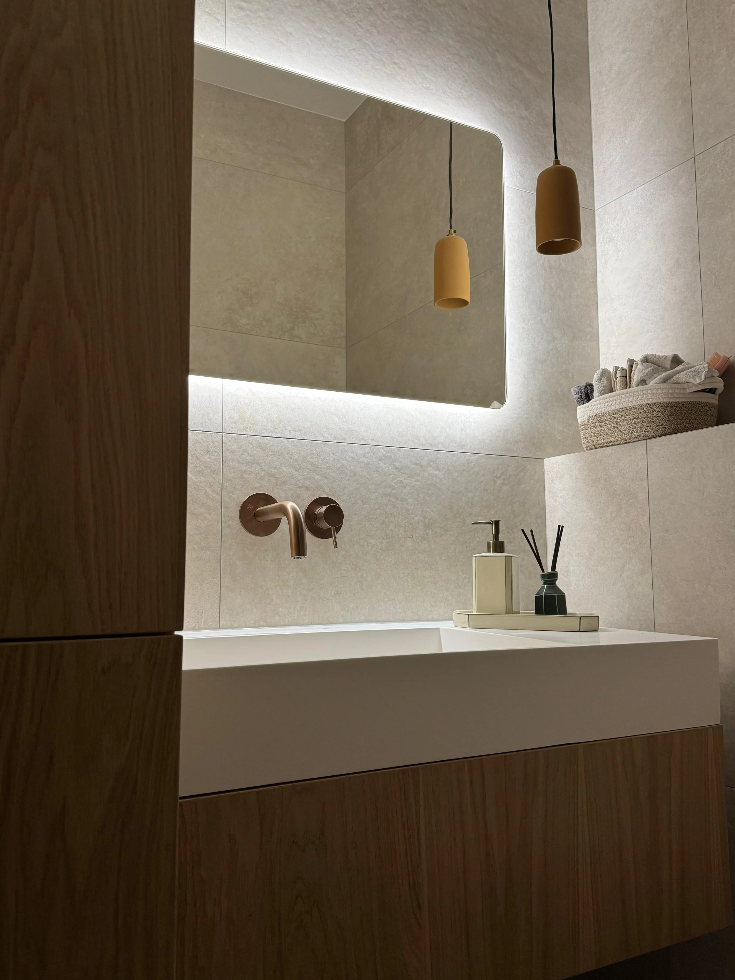 Modern bathroom vanity with a white rectangular sink, wall-mounted bronze faucet, a large backlit mirror, tan hanging lights, a container with rolled towels, and reed diffusers on a tray.