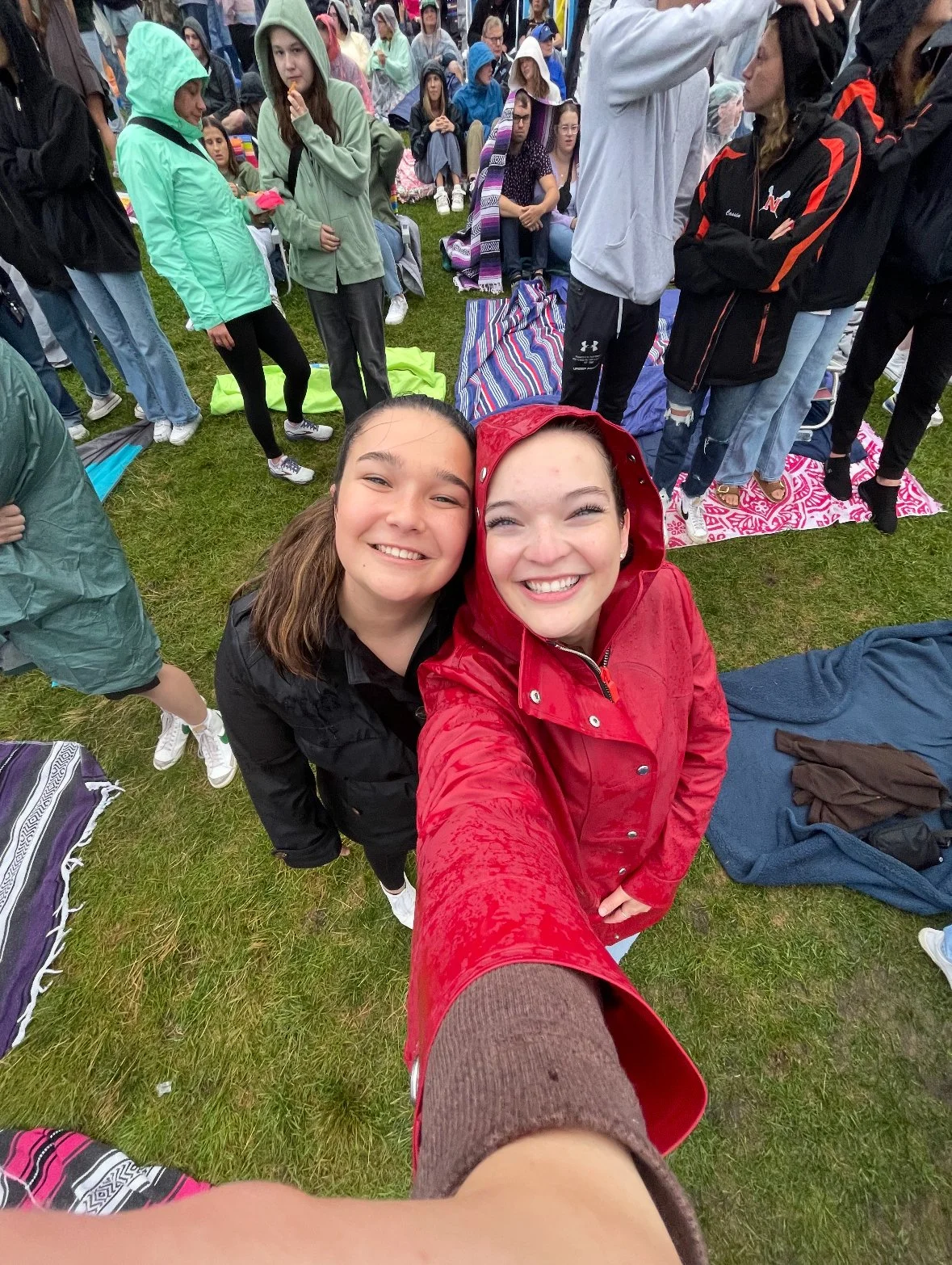 Two smiling young women taking a selfie at an outdoor event in the rain. One is wearing a red raincoat and the other a black jacket. Many people in rain gear are gathered on a grassy field behind them.