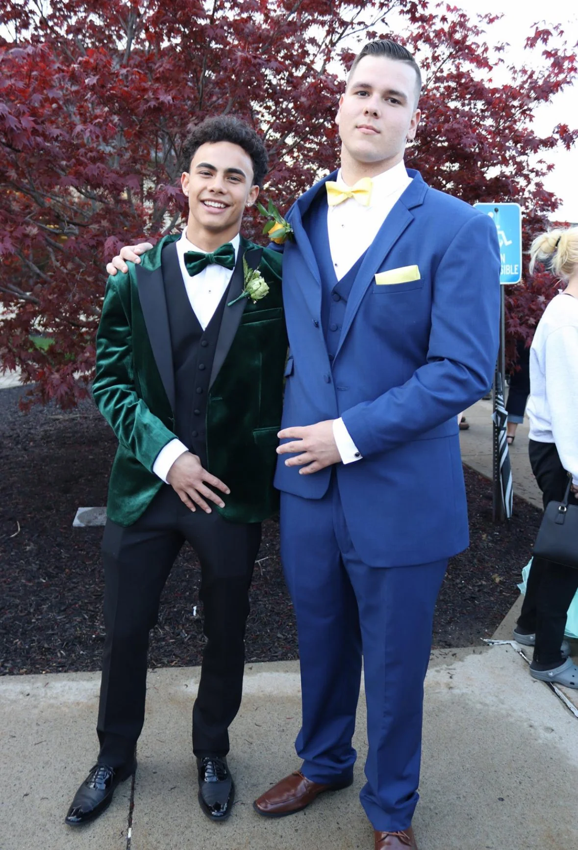 Two young men dressed in formal suits, one in a green velvet blazer and the other in a blue suit, standing outdoors in front of a red tree with other people in the background.