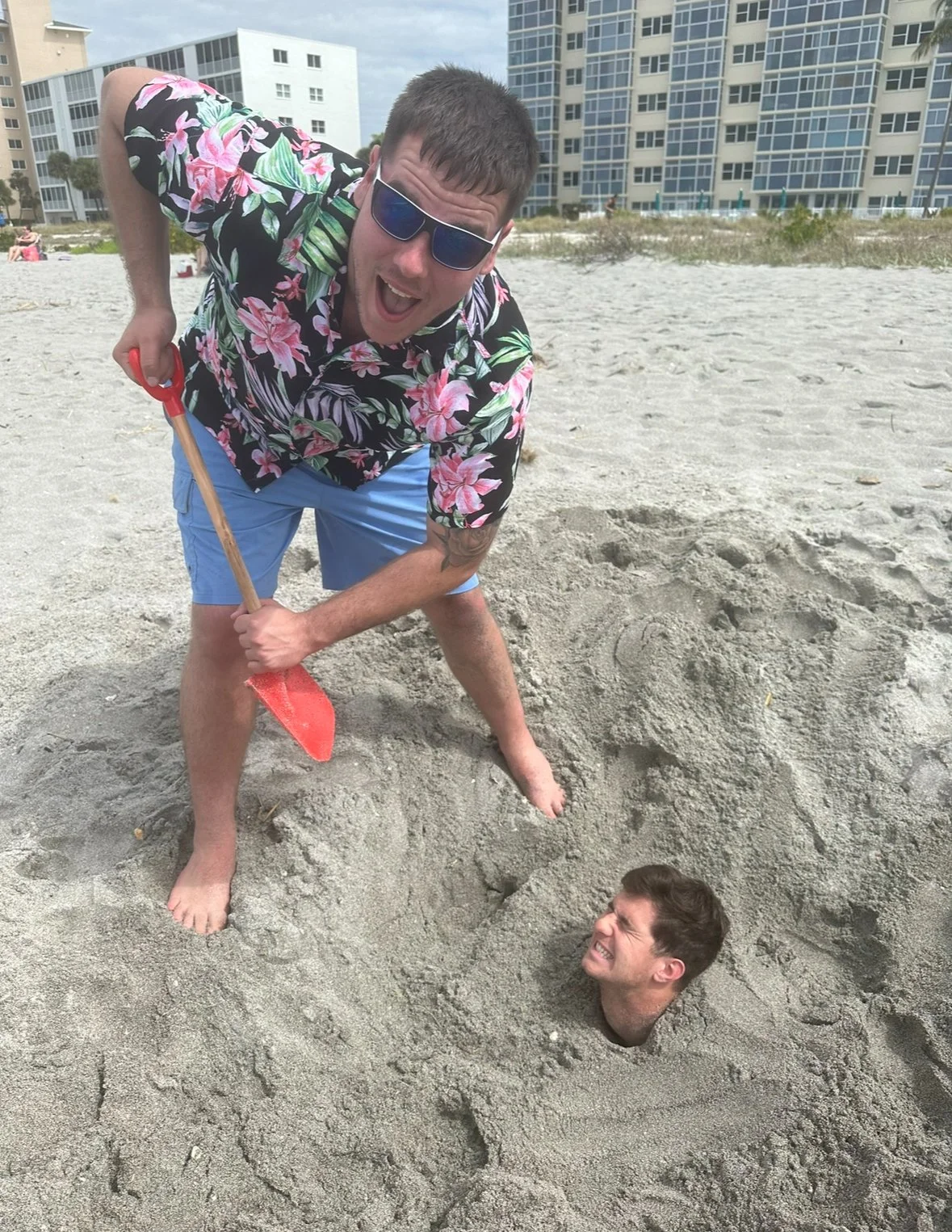 Two men at the beach. One man is standing in a hole, holding a small shovel, while the other man has his head buried in the sand, with only his head visible. There are high-rise buildings in the background.
