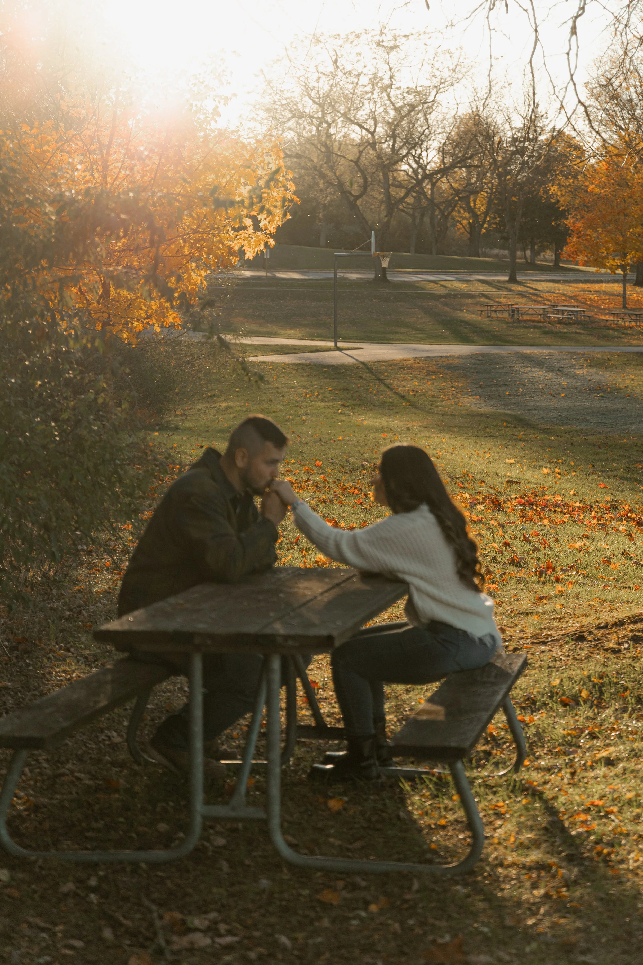 A man and woman sitting at a picnic table in a park during autumn, with a basketball hoop and trees in the background. The woman is holding the man's hand, and they are facing each other.