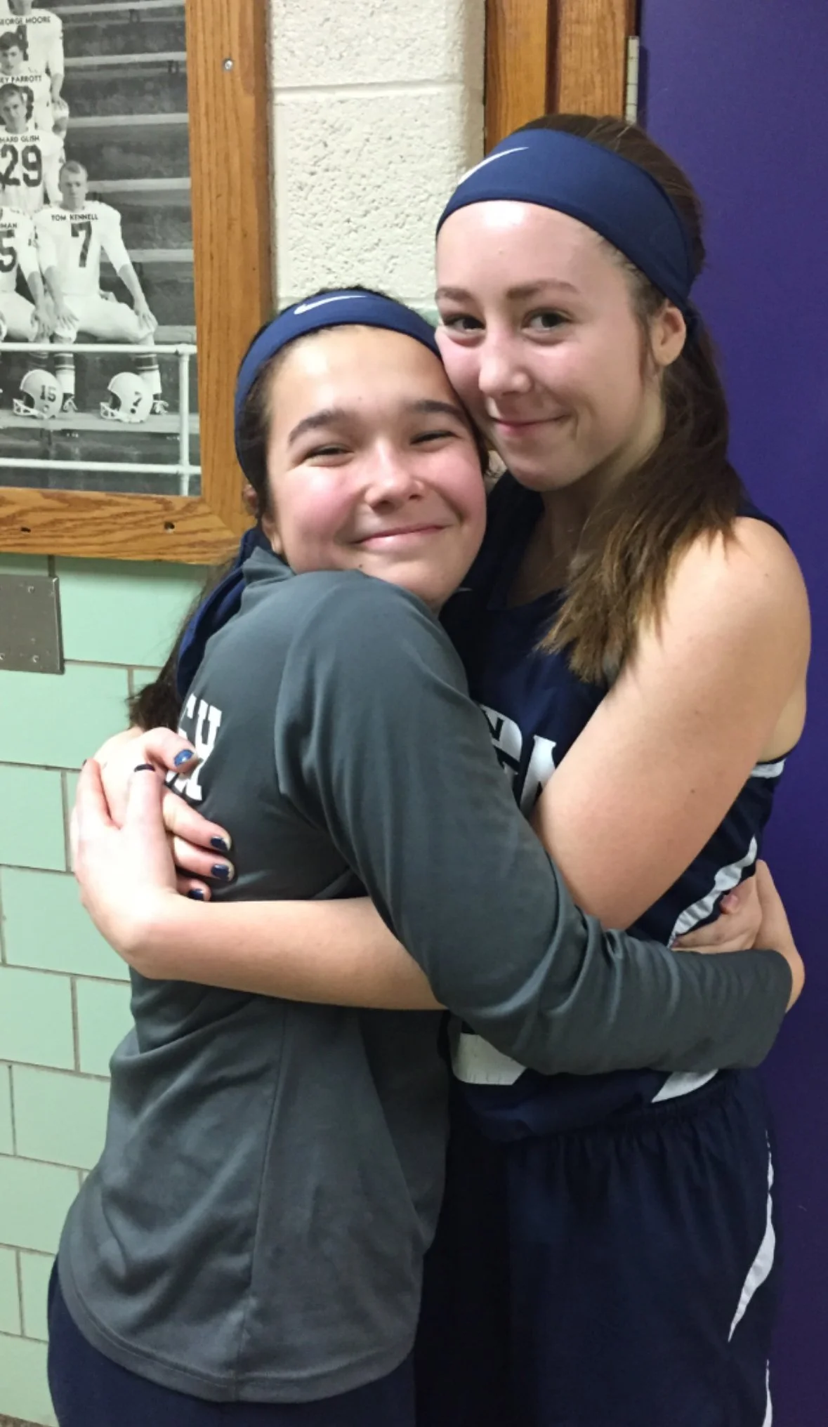 Two girls in athletic uniforms hugging and smiling at the camera in an indoor sports setting.