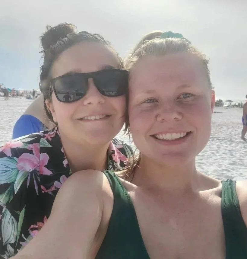 Two women taking a selfie at the beach with sand and people in the background. One woman is wearing sunglasses and a floral shirt, the other has a green tank top.