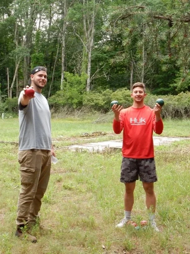 Two young men standing outdoors in a grassy area with trees in the background, each holding a kettlebell.