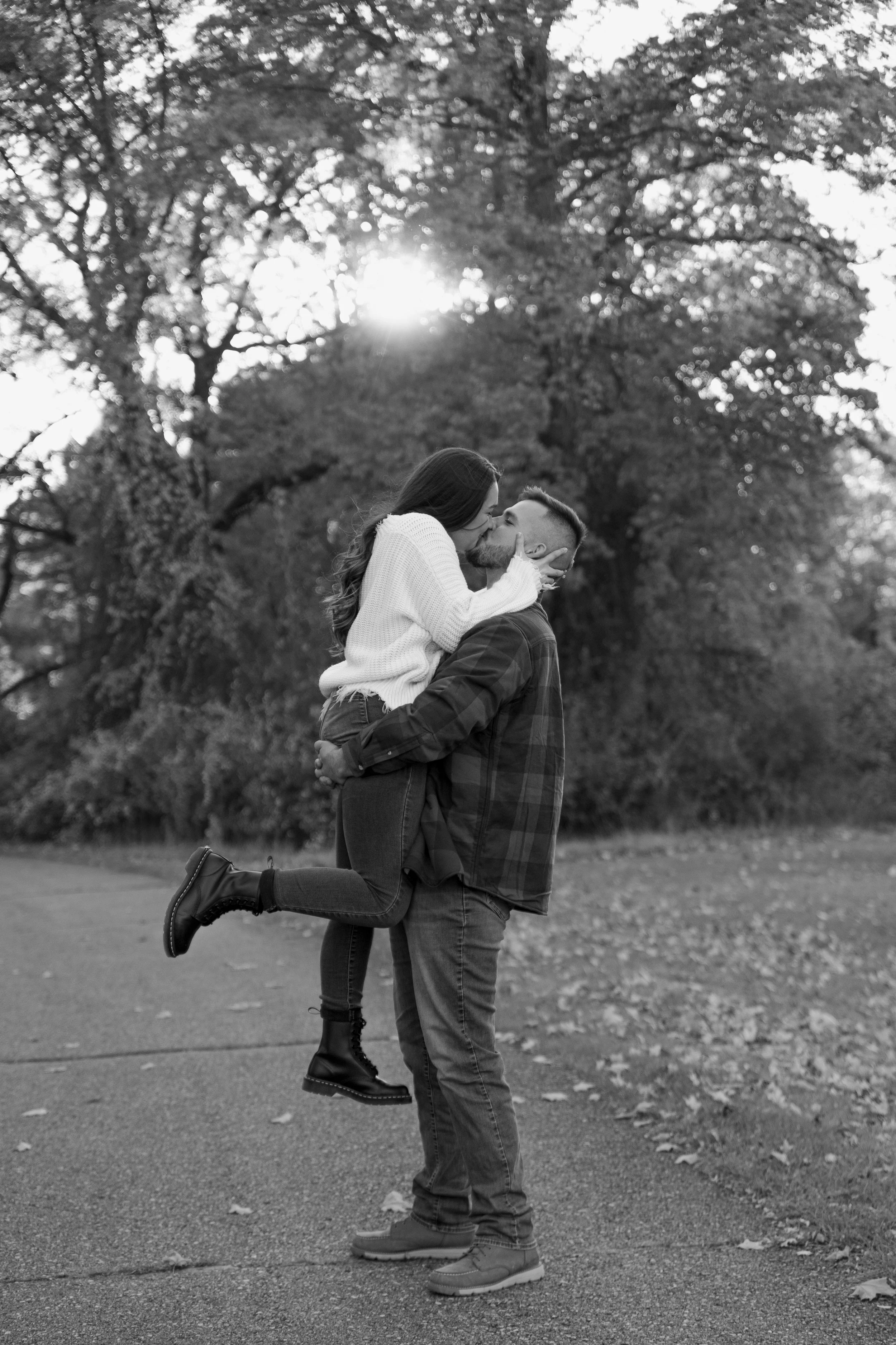 A black and white photo of a couple outdoors, with the man lifting and holding the woman as they kiss. The woman wears a white sweater and boots, and the man wears a plaid shirt and jeans, surrounded by trees and fallen autumn leaves.