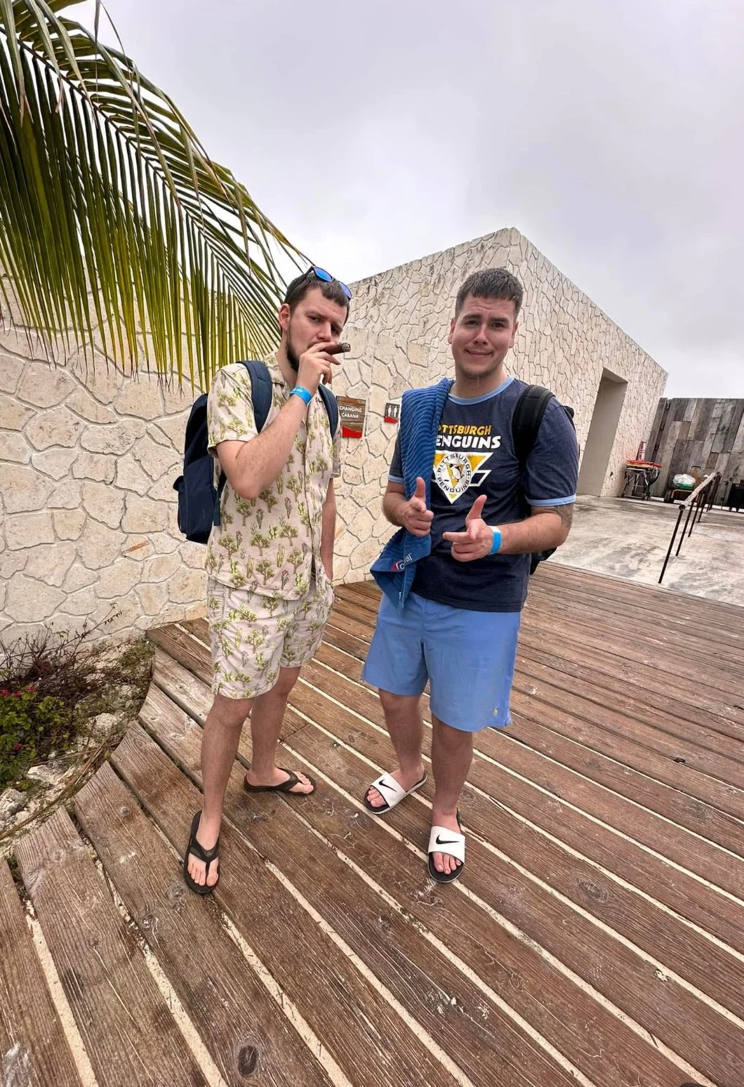 Two young men standing on a wooden deck outdoors under an overcast sky, one wearing a tropical shirt and shorts, the other wearing a Pittsburgh Penguins t-shirt and shorts, both with backpacks and sandals, near a stone wall with tropical plants and signage in the background.