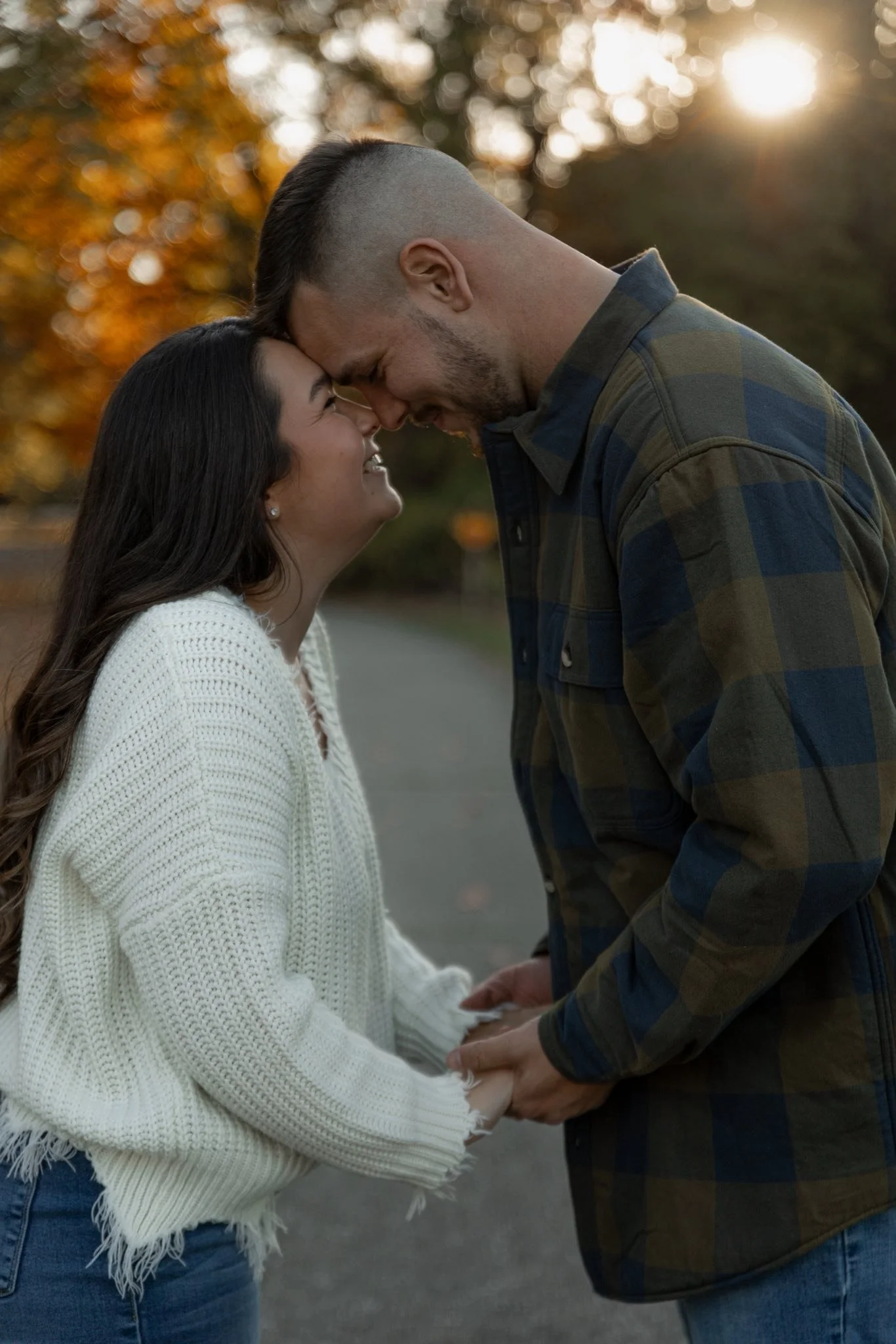 A couple holding hands and smiling, with their foreheads touching in an outdoor setting during sunset, with fall-colored trees in the background.