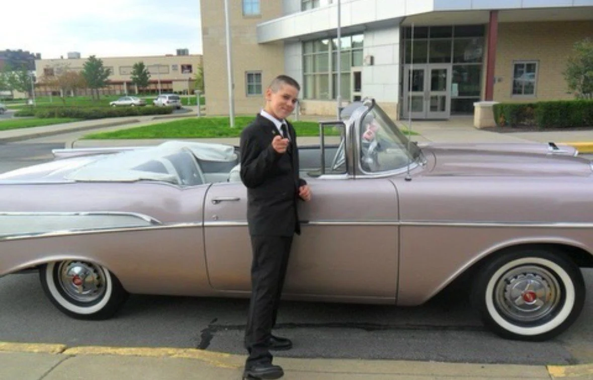 Young man in a black suit standing next to a vintage silver convertible car outside a modern building